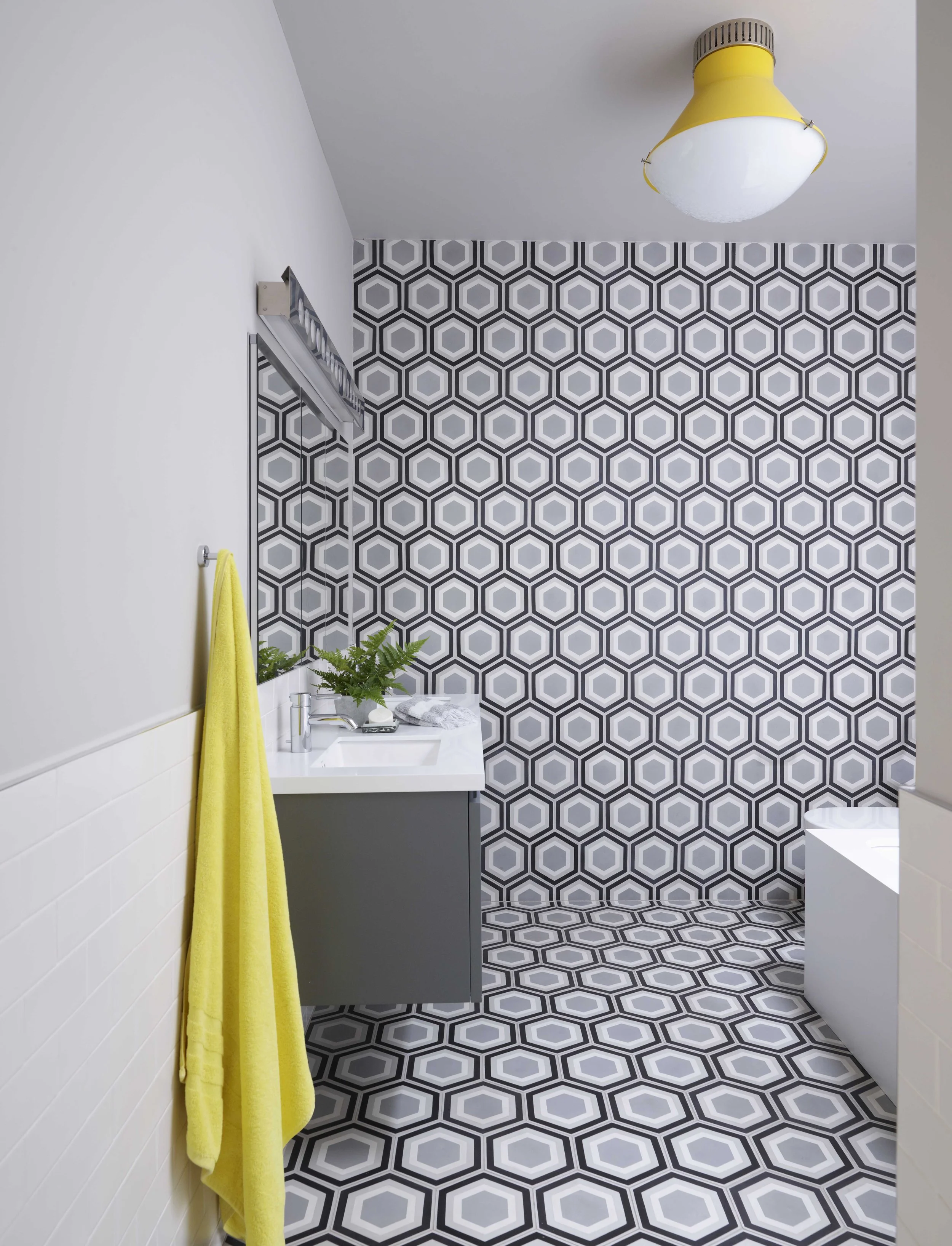A modern bathroom with black, gray, and white hexagonal patterned tiles on the wall and floor, a gray vanity with a white sink, a yellow towel hanging on the wall, and a ceiling light fixture.