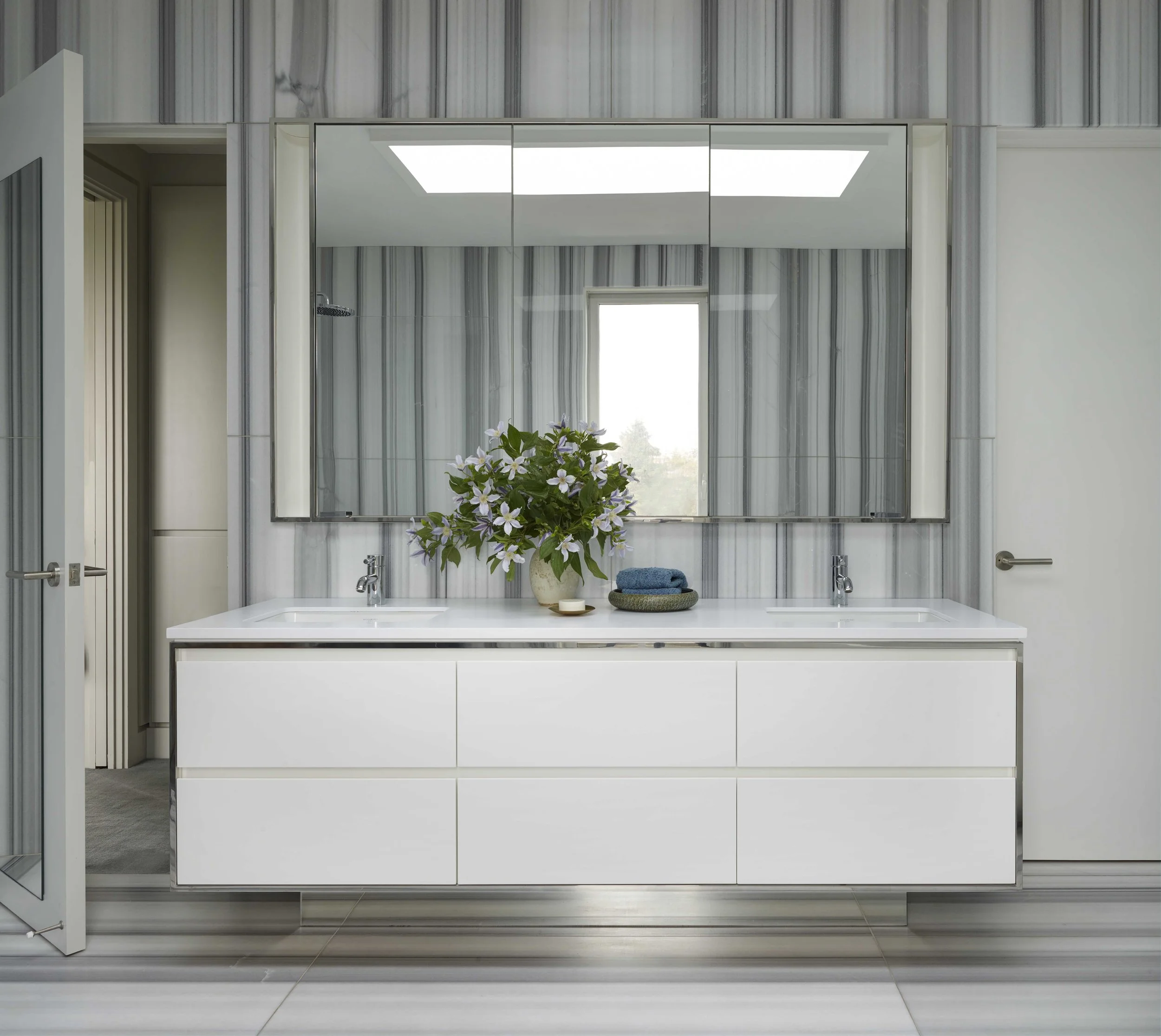 Modern bathroom vanity with white drawers, large mirror with 3 panels, gray marble wall, window with view outside, vase with flowers, and towels.