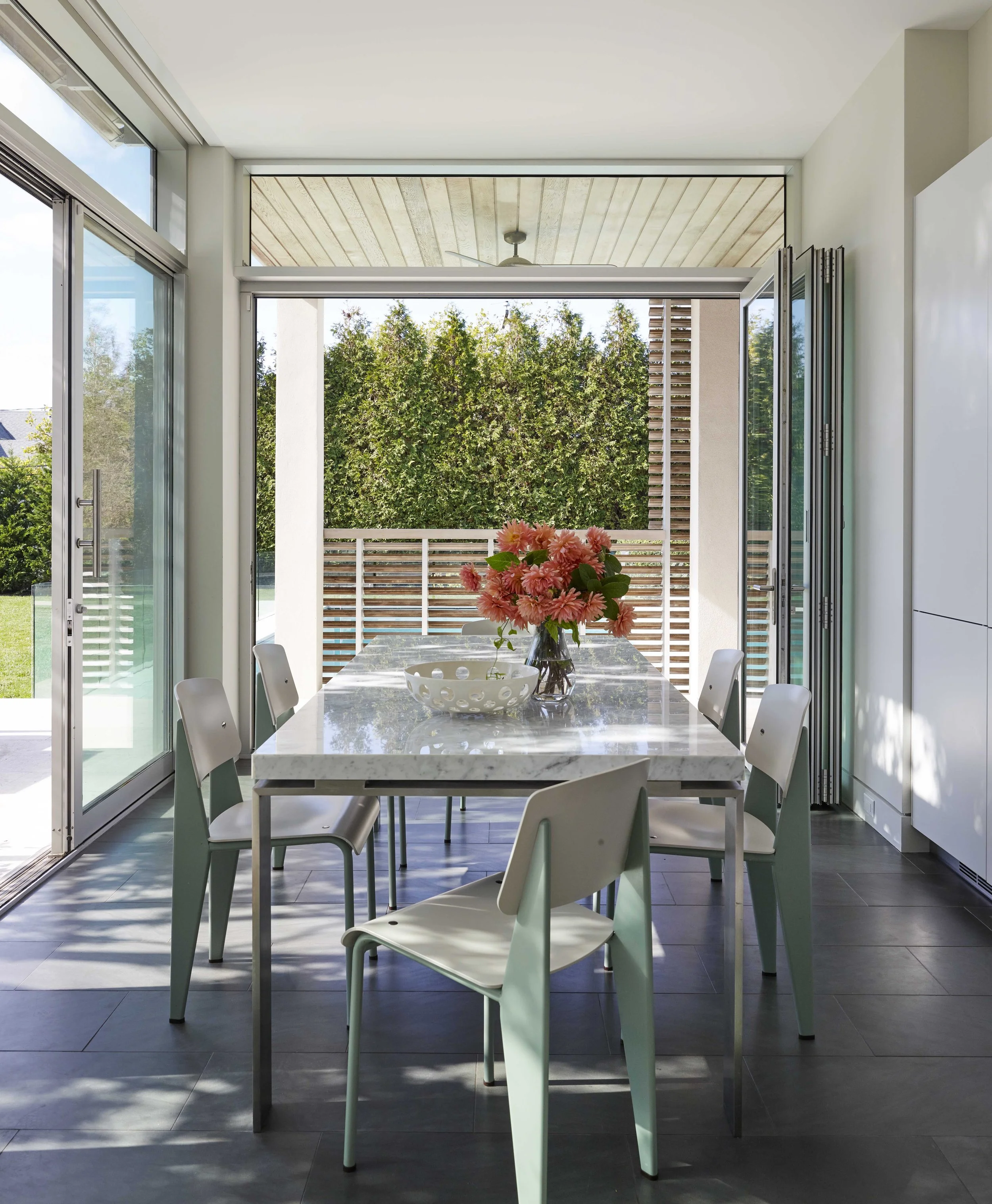 A modern dining area with a marble table, six white chairs, a flower arrangement, and a large open door leading to a patio with a green yard and trees.
