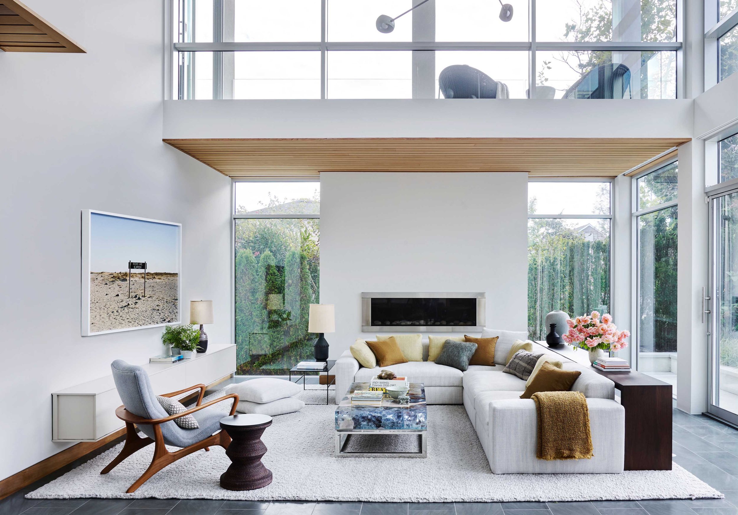 Modern living room with large windows, white walls, a sectional sofa with pillows, a glass coffee table, a mid-century armchair, and a balcony above.