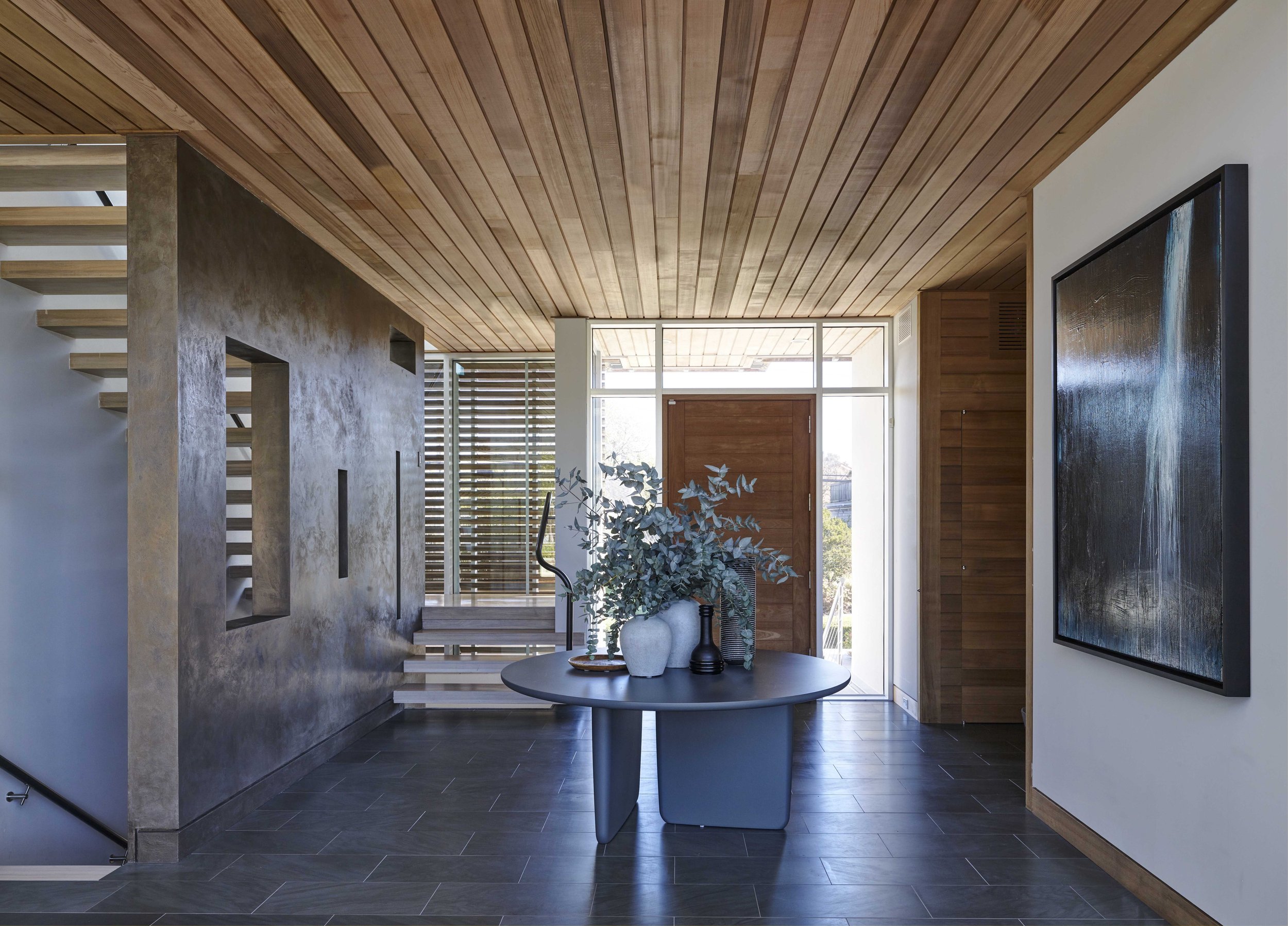 Modern entryway with wooden ceiling, stairs, and botanical arrangement on a dark table.