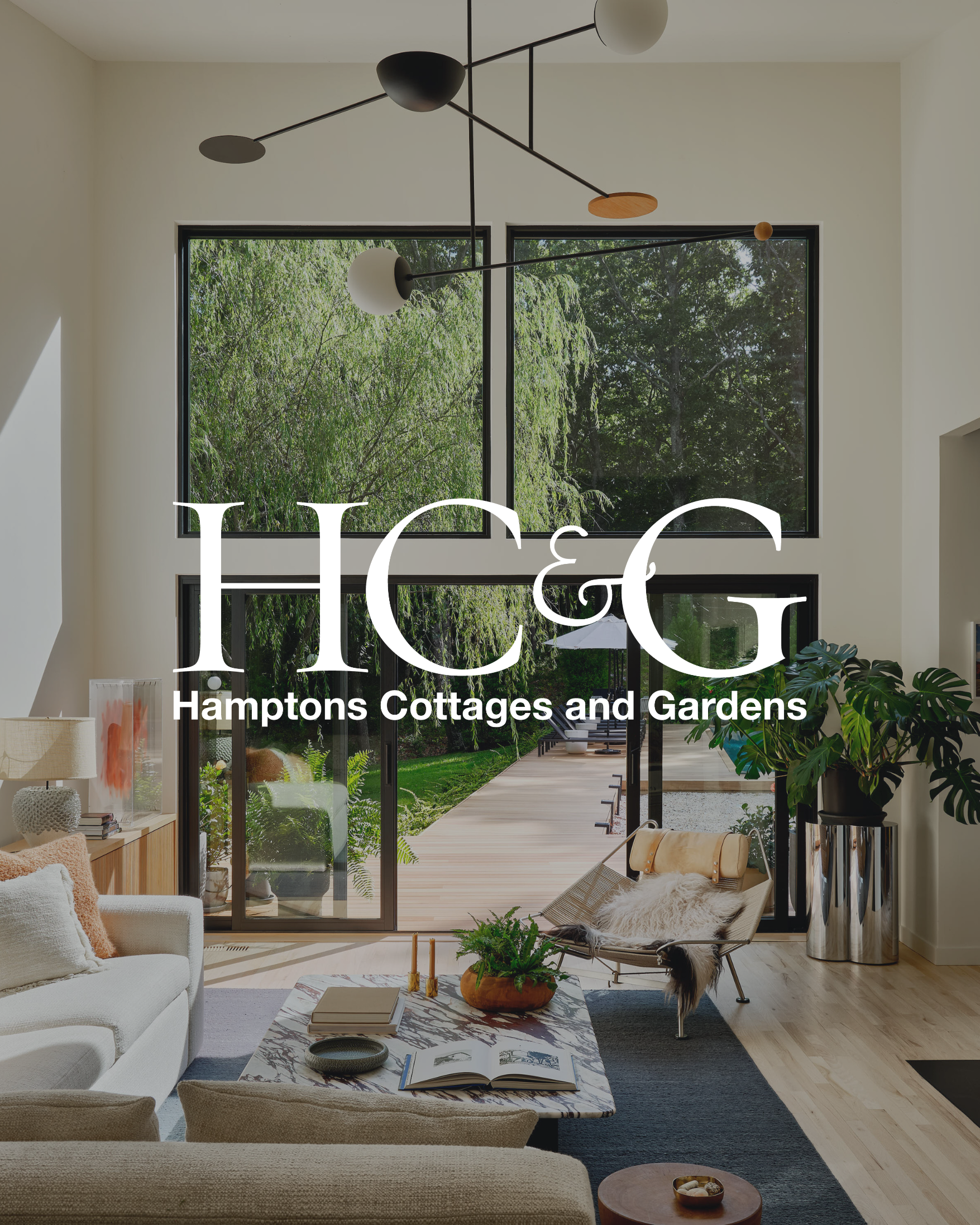 Modern living room with large windows revealing a green backyard, a white sofa, a marble coffee table with books and a plant, a side table with a lamp, a lounge chair with a throw blanket, and a large potted plant. Overlaid text reads 'HCG Hamptons Cottages and Gardens.'