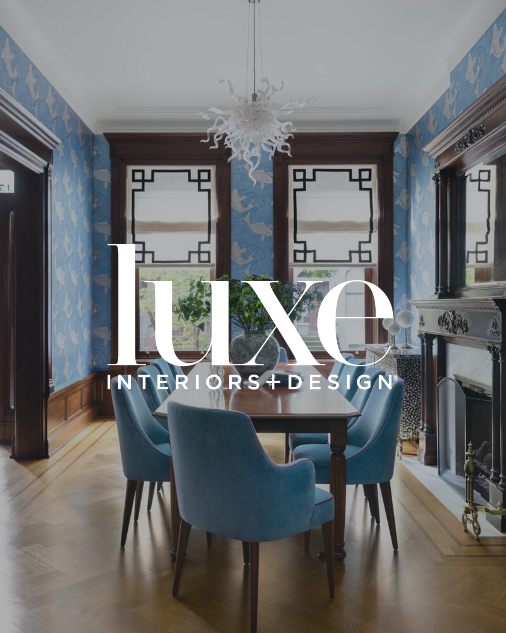 Interior dining room with a wooden table and blue chairs, blue patterned wallpaper, large windows with black geometric trim, a fireplace with a decorative mirror, and a white chandelier hanging from the ceiling.