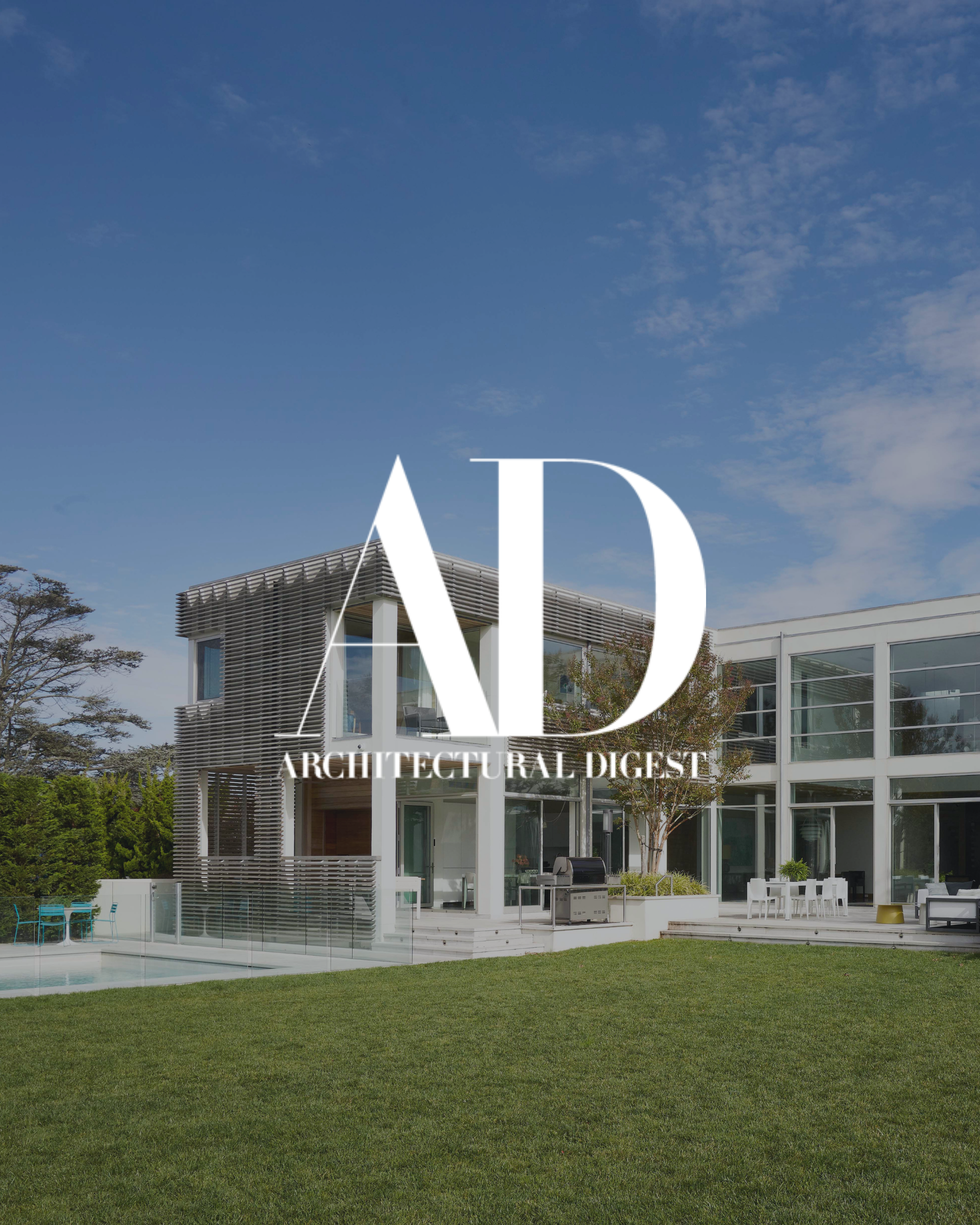 Modern house with large glass windows and a patio area, set against a blue sky with some clouds, surrounded by greenery.