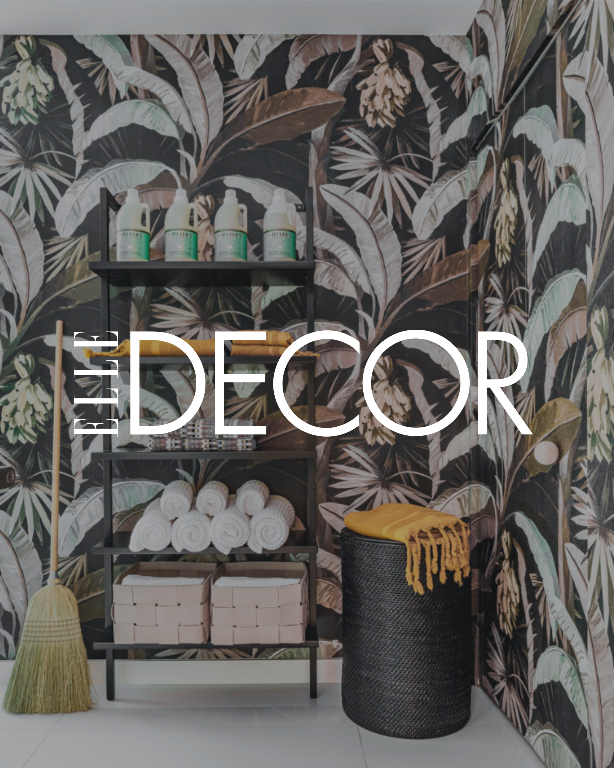 A decorative room with dark tropical leaf wallpaper, a black shelving unit holding white towels and bottles, a black basket with a yellow blanket, and a gold broom leaning against the wall.