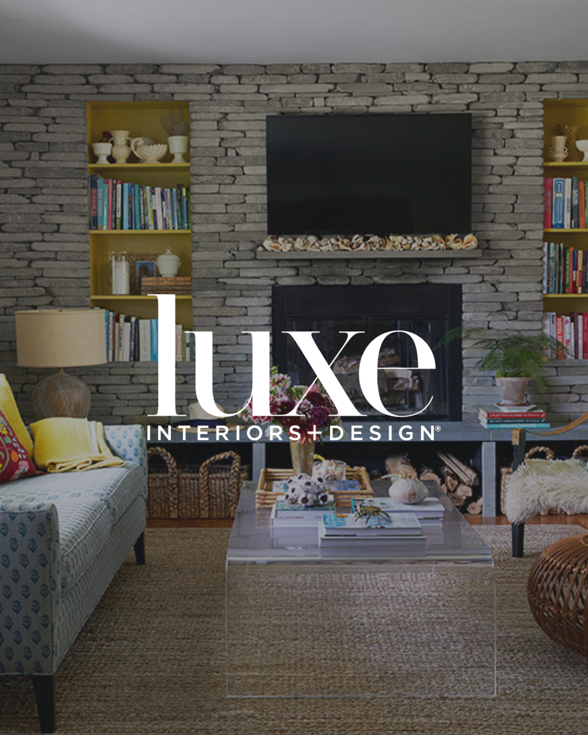 Living room with a brick fireplace, a flat-screen TV above it, yellow bookshelves on each side, a patterned sofa with colorful pillows, a glass coffee table with books and flowers, a side table with a plant, and a woven ottoman.