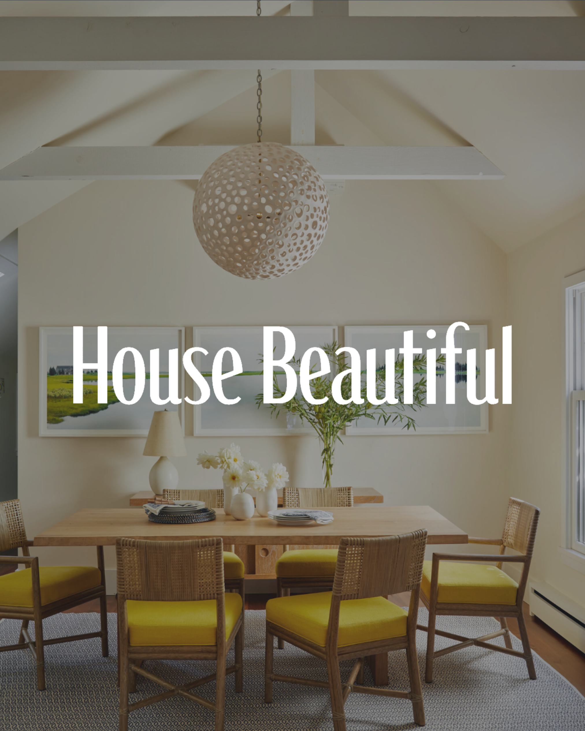 A dining room with a wooden table, yellow cushioned chairs, a white vase with flowers, a lamp, and three framed landscape pictures on the wall. A spherical decorative light fixture hangs from the ceiling.