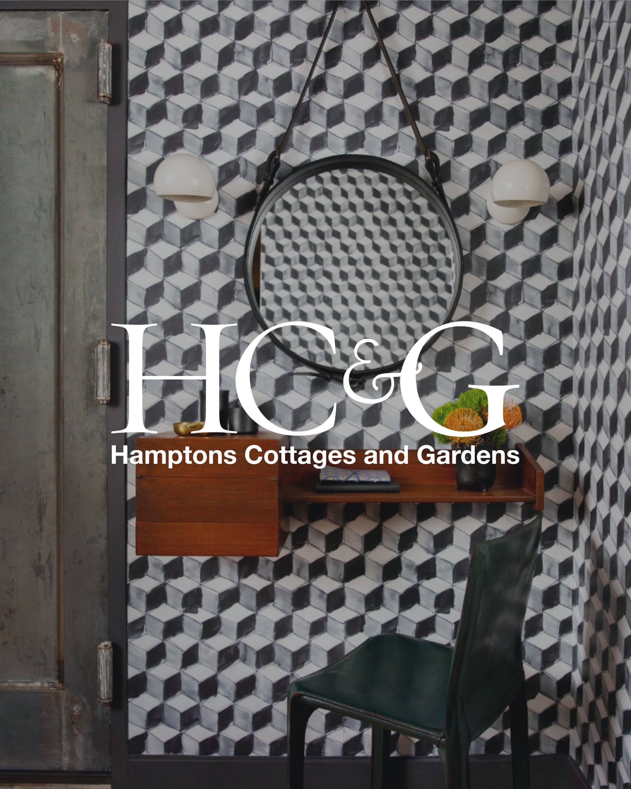 Decorative interior space with black and white 3D geometric patterned wallpaper, a round mirror, two white wall sconces, a small wooden desk with a flowerpot and some books, and a green leather chair.