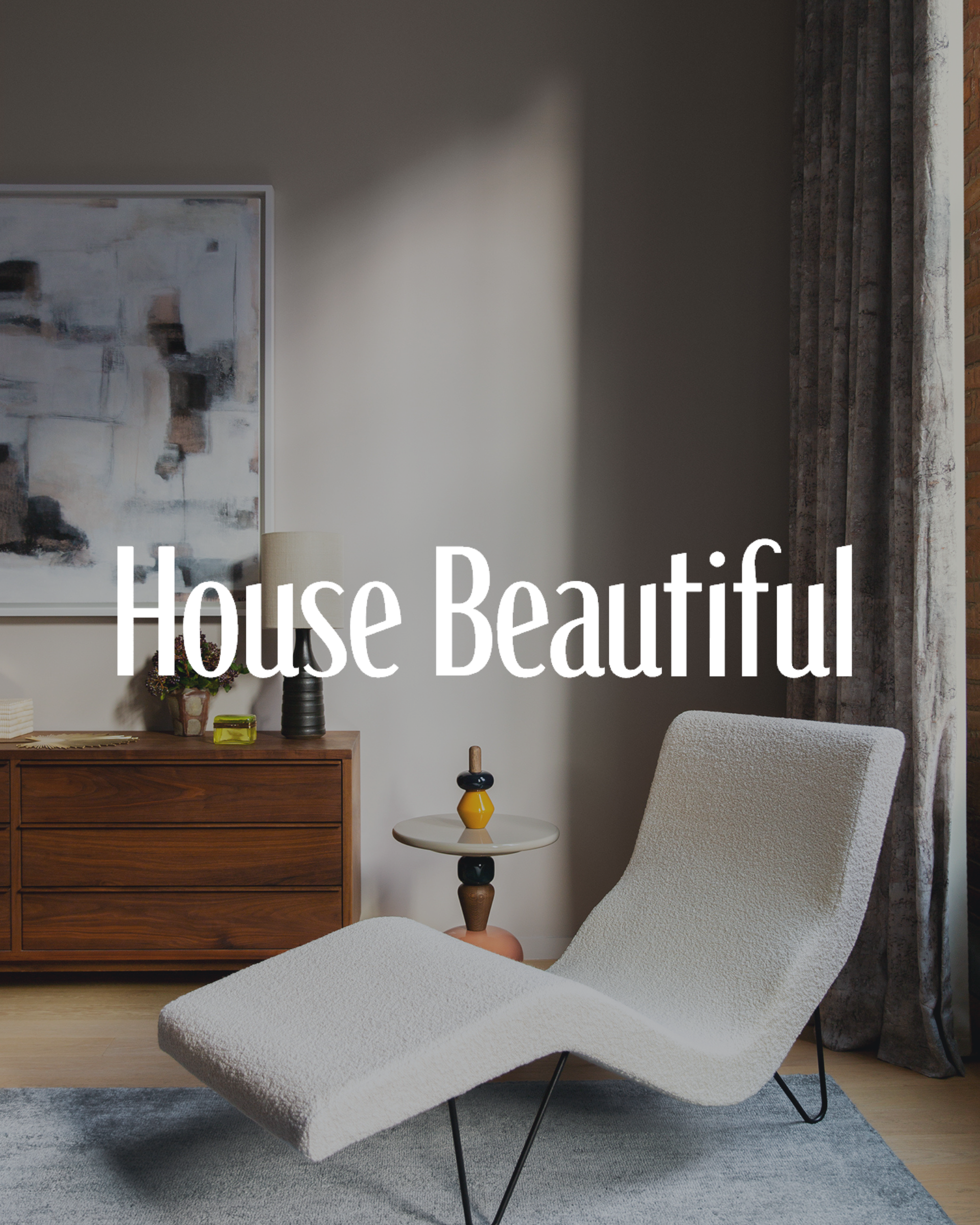 Modern living room with a white lounge chair, wooden console, abstract artwork, small side table, decorated with a vase and small decor items, with a curtain and brick wall in the background.