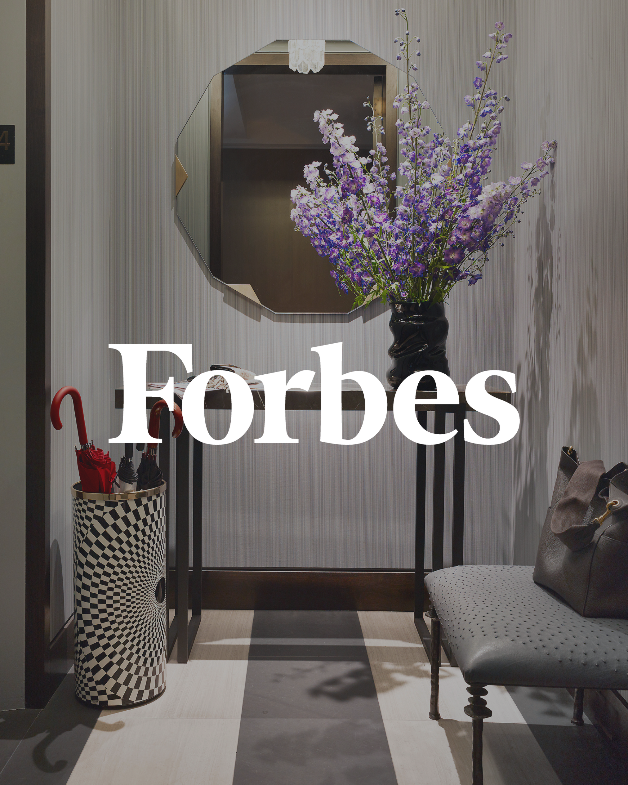 A hallway with a large mirror on the wall, a vase of purple flowers on a small table, a bench with a handbag, and an umbrella stand with umbrellas, with the Forbes logo overlaid.