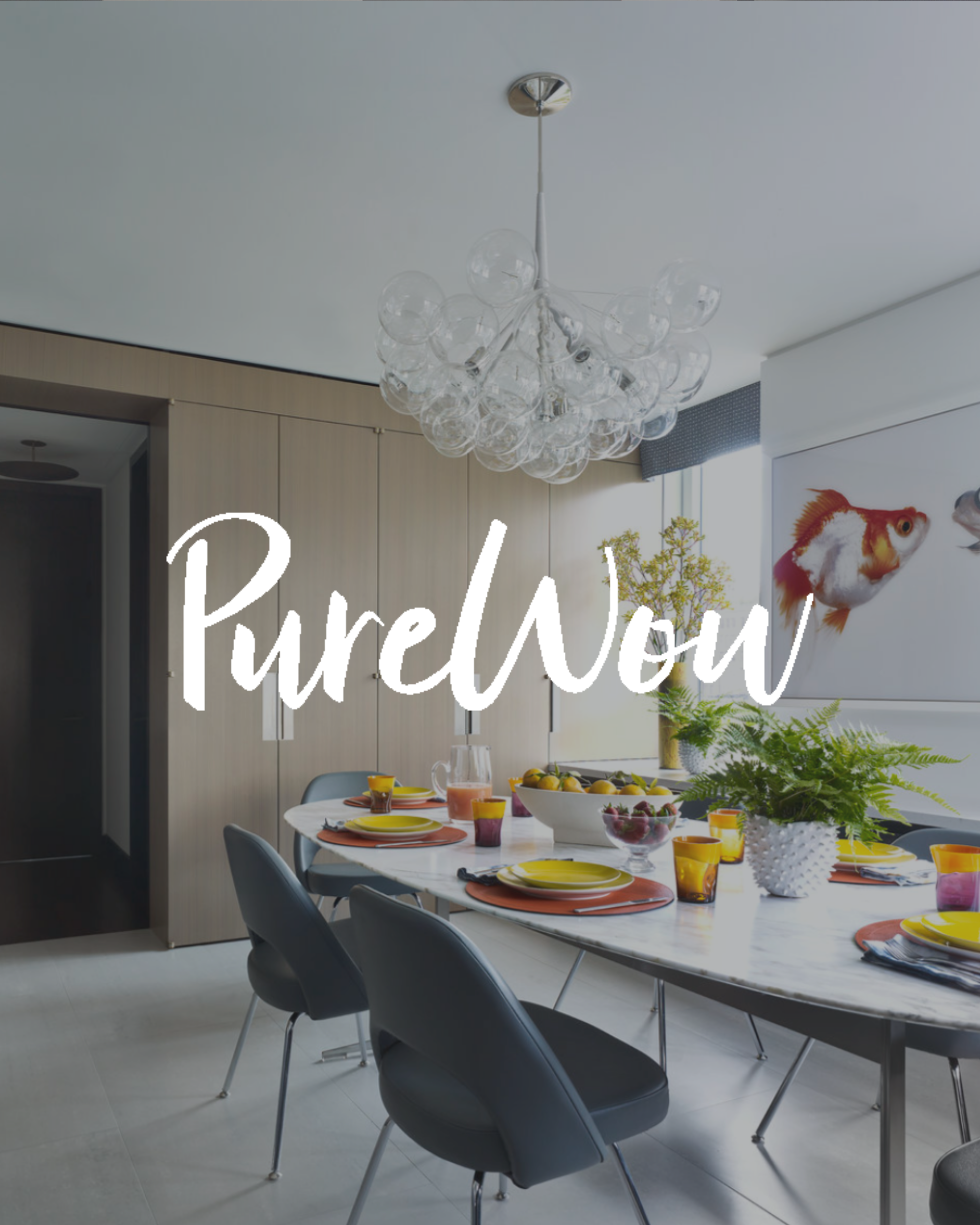 Modern dining room with a white marble table set with yellow and purple plates, glasses, and bowls of fruit, surrounded by gray chairs, with a chandelier hanging from the ceiling, large window, and wall art of a fish.