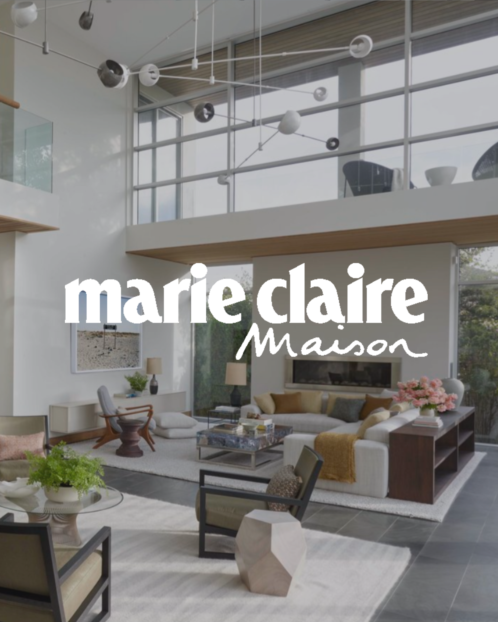 Modern living room with large windows, white walls, and contemporary furniture, including a sectional sofa, armchair, coffee table, and decorative items. The logo of Marie Claire Maison is prominently displayed across the center.