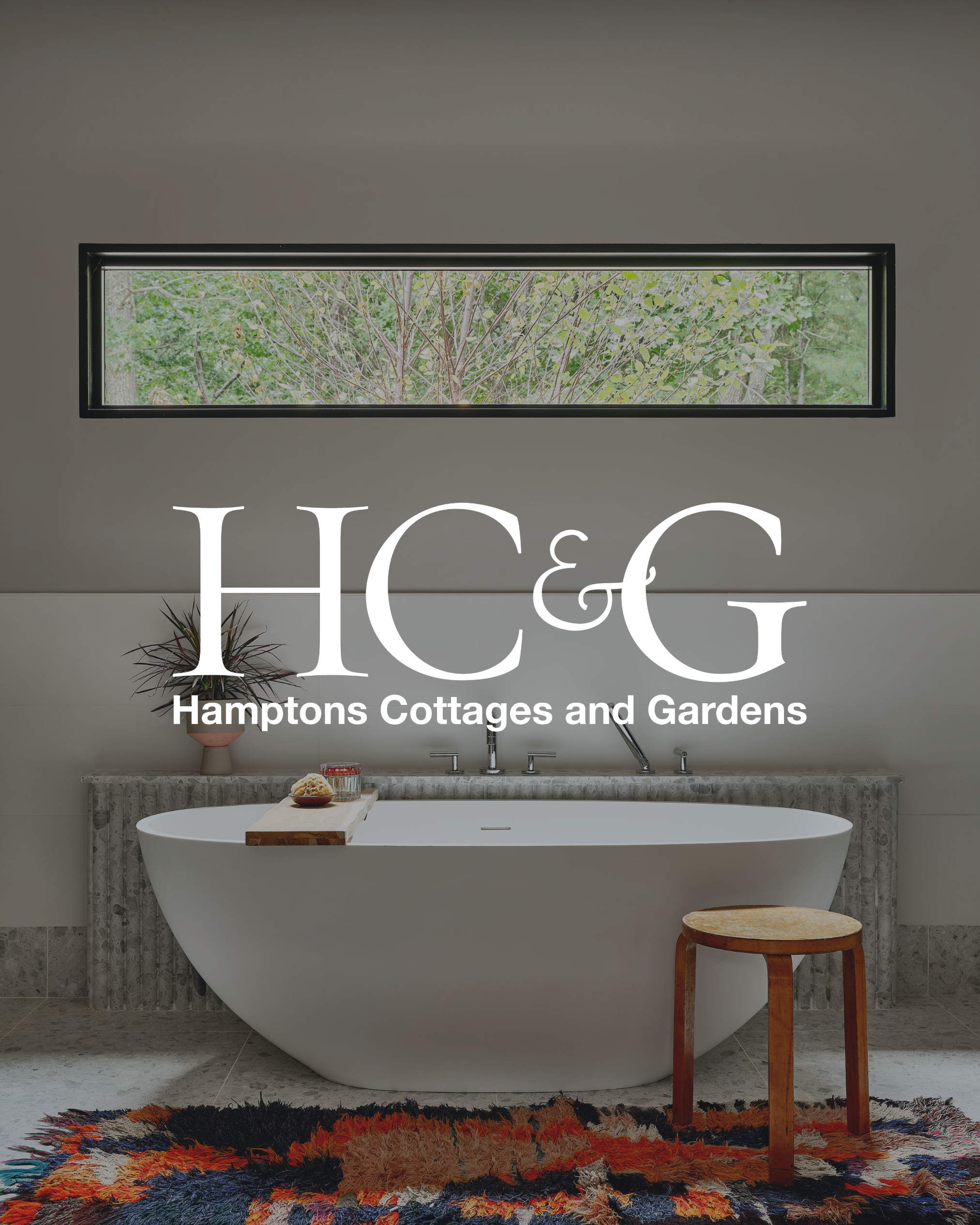 Modern bathroom with a freestanding bathtub, wooden stool, colorful rug, and a long horizontal window showing greenery outside.