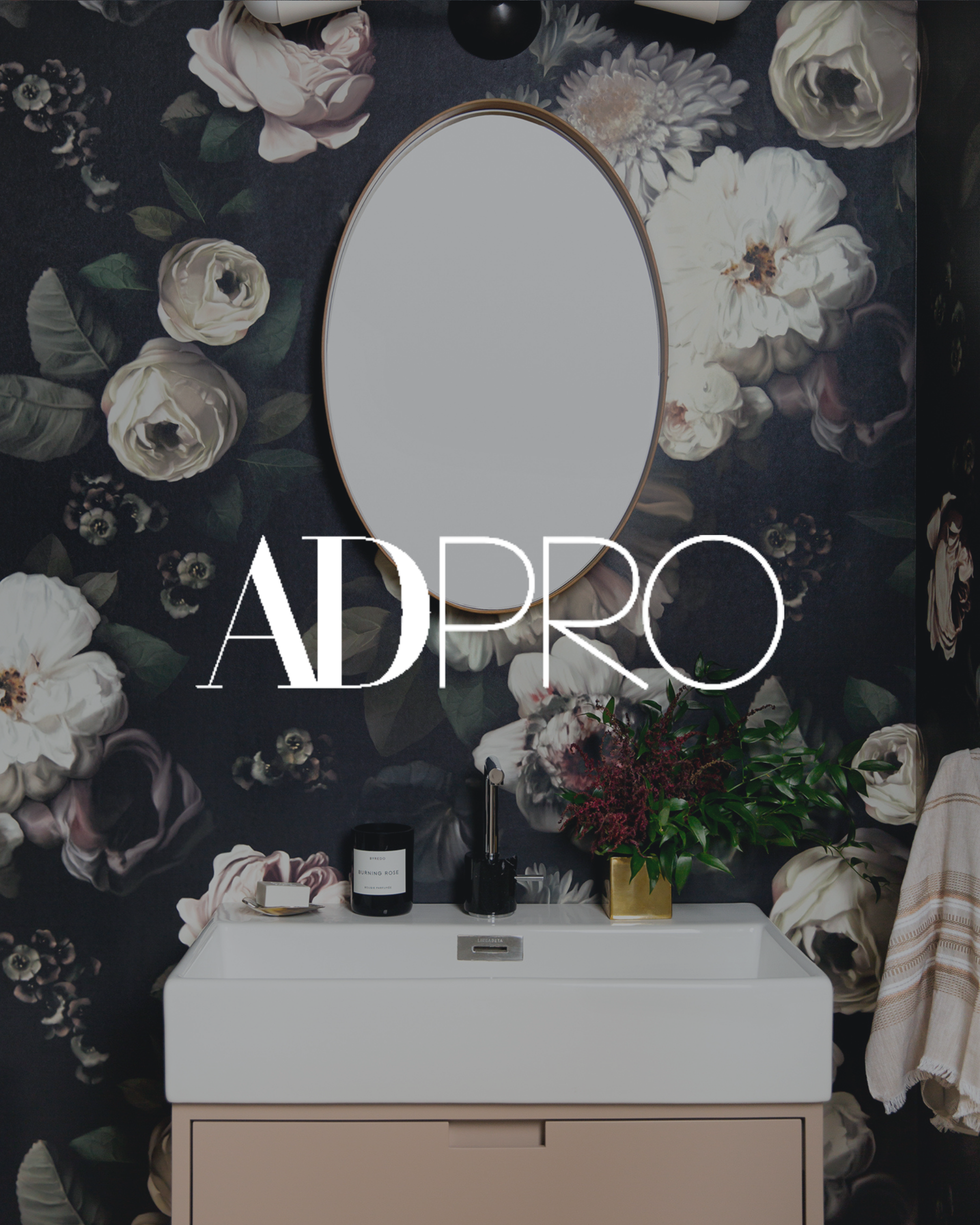 A bathroom vanity with a white sink, black soap dispenser, black candle, and a gold vase with green and purple flowers against a floral wallpaper background with a round mirror.