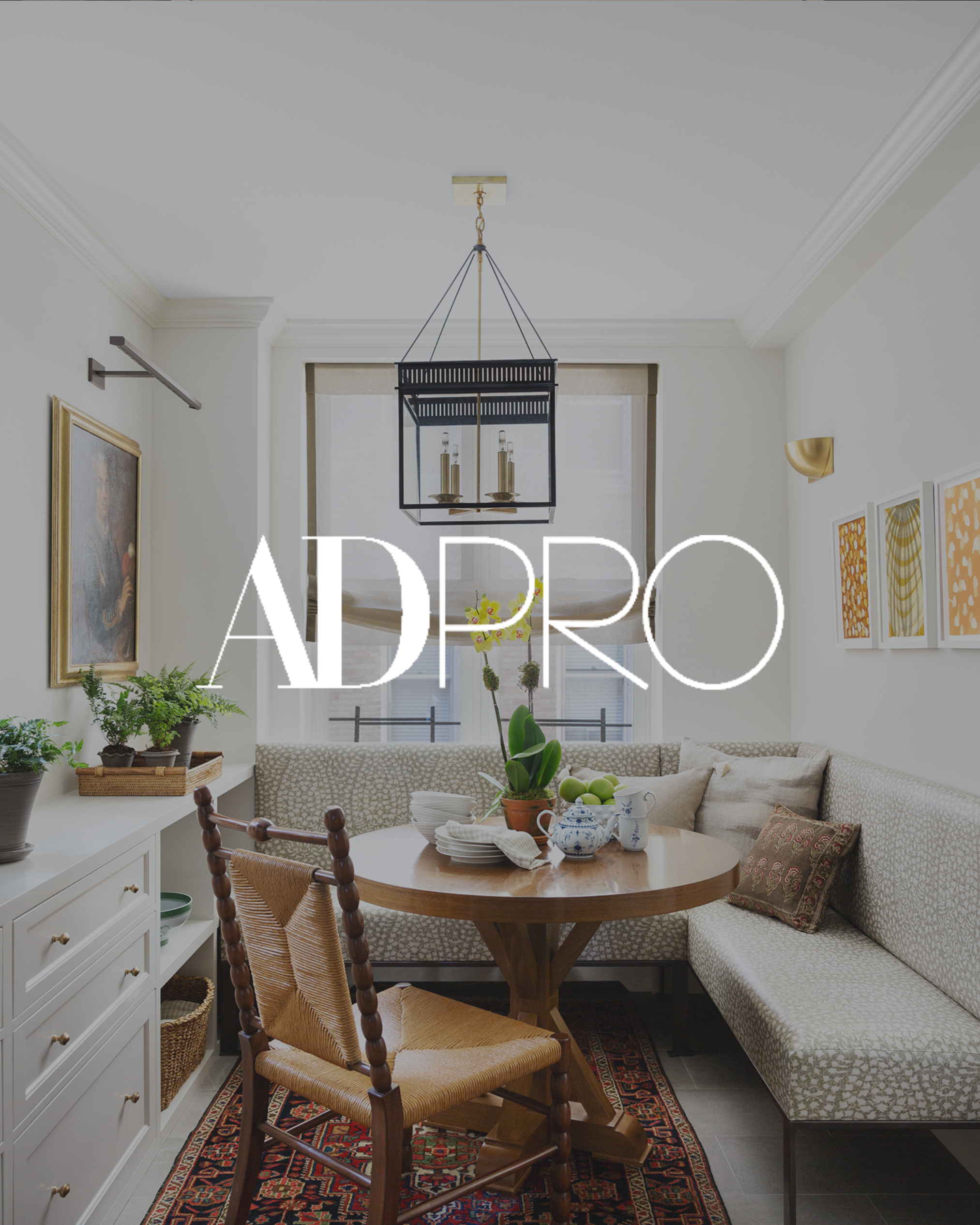 Bright dining area with a wooden round table, cushioned bench seats, potted plants, and a black metal chandelier hanging from the ceiling.