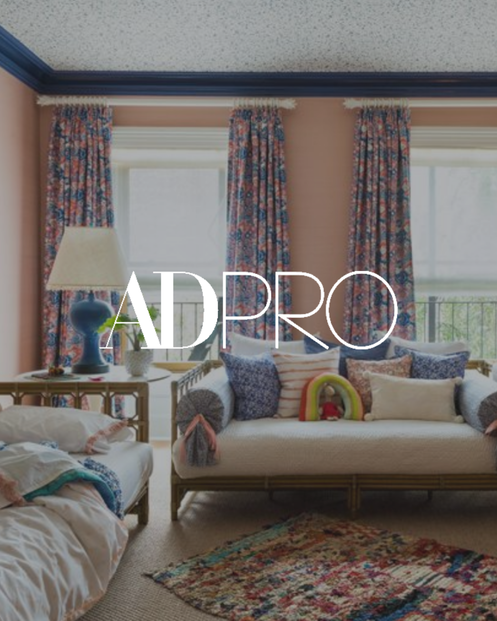 Living room with a sofa filled with colorful pillows, a side table with a lamp and potted plant, large windows with floral curtains, and an area rug on the carpeted floor.