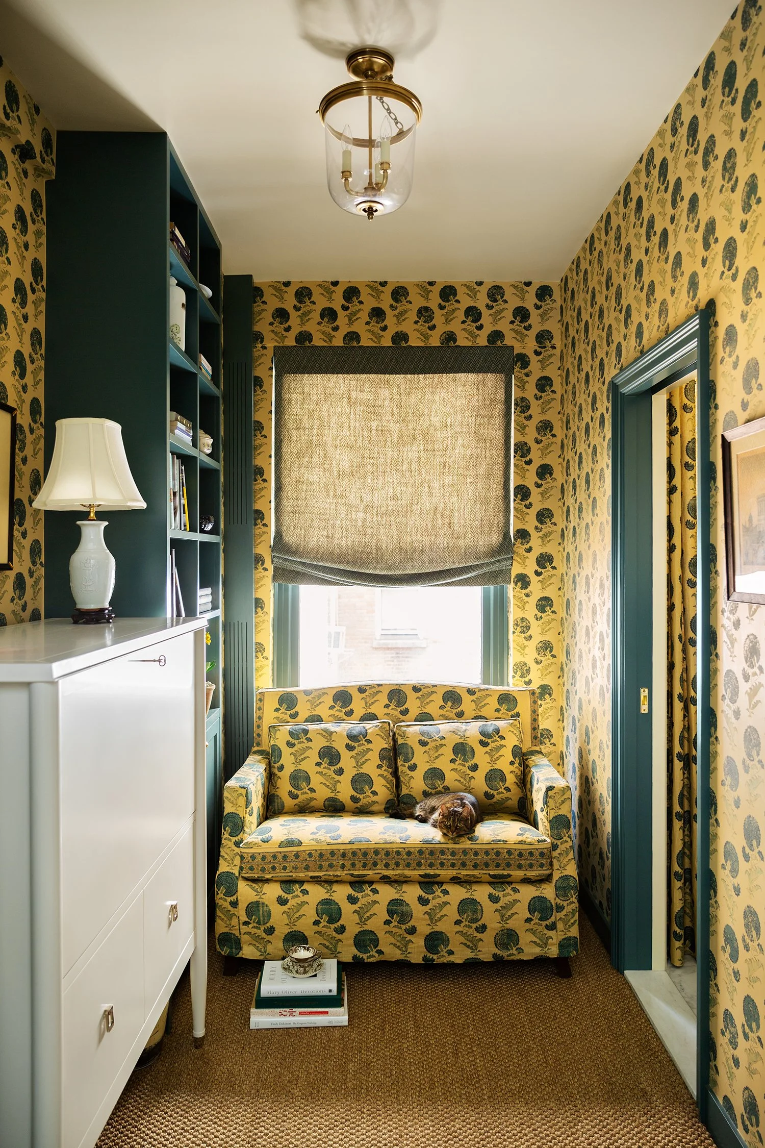 A small room with floral-patterned wallpaper and matching upholstery on a loveseat, a window with a roman shade, a white cabinet with a lamp on top, a bookshelf filled with books, a small cat resting on the loveseat, and a stack of books on the carpe
