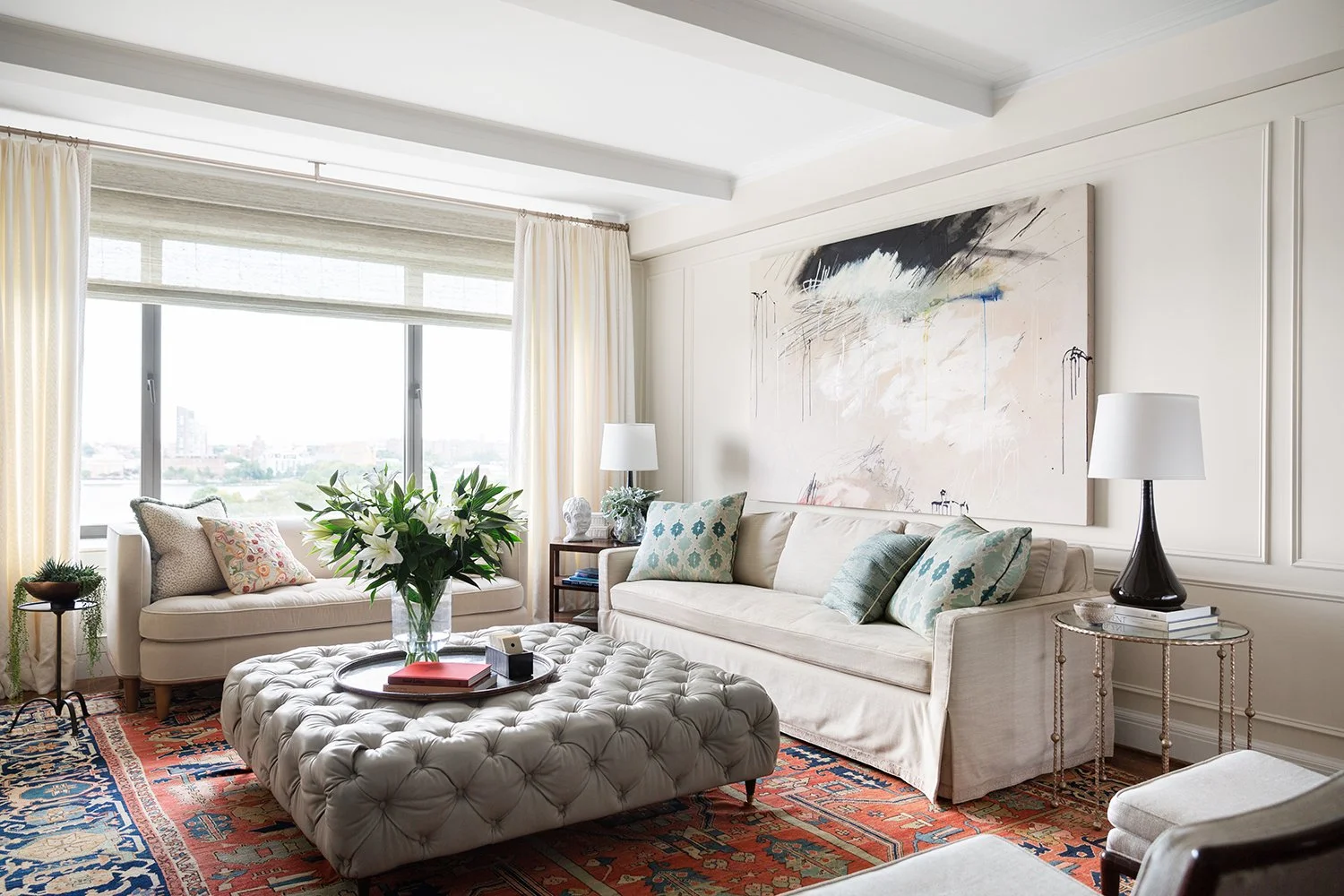 Living room with a large beige sofa, a tufted ottoman coffee table with a flower vase, a side table with a lamp, and an abstract wall art in the background. There are decorative cushions on the sofa, and a large window with curtains letting in natura