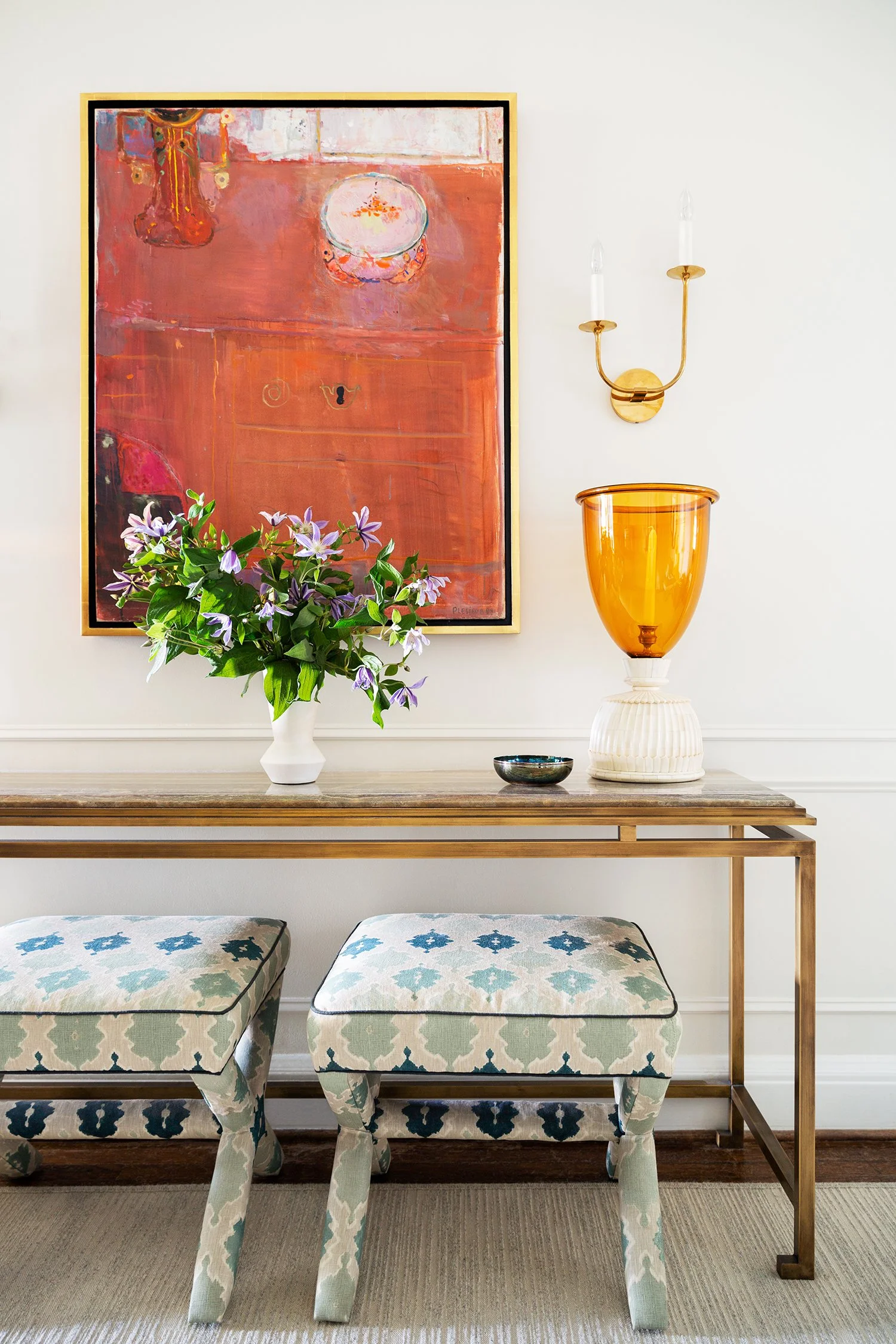 Interior decor of a living space with a wooden console table, floral arrangement in a white vase, a large amber glass vase on a white base, abstract artwork on the wall, a gold wall sconce with two light bulbs, and two patterned upholstered stools.