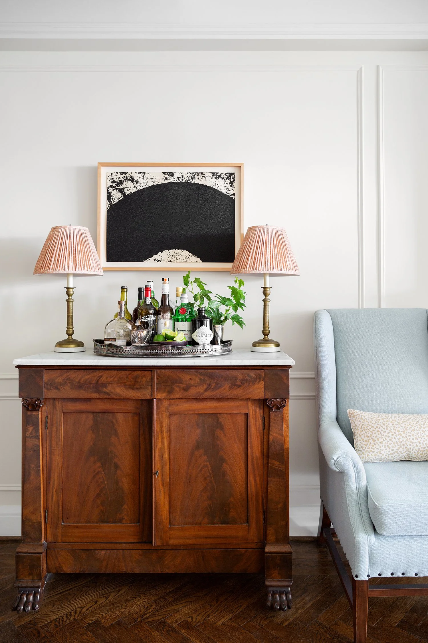 A wooden sideboard with a marble top, holding a silver tray with assorted liquor bottles, lime slices, and a potted plant. Two pink lamps with brass bases flank the sideboard. Above, a black and white abstract painting hangs on the white wall. Part o