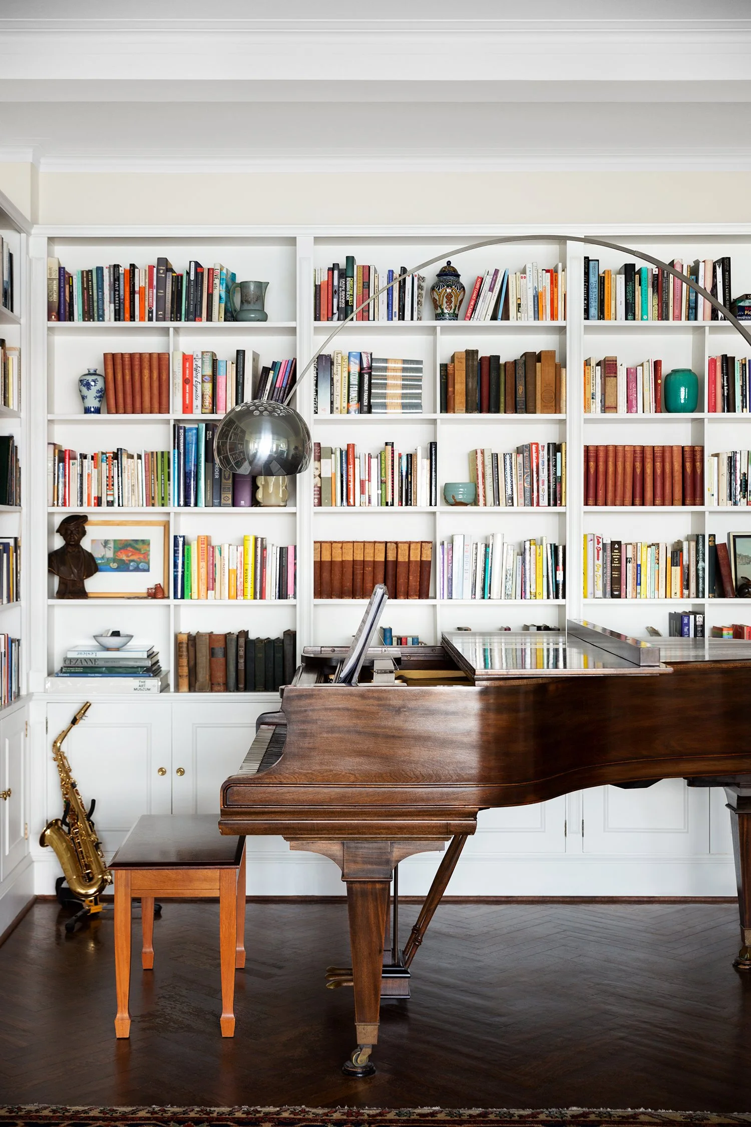 A wooden grand piano with a small bench in front of a white bookshelf filled with colorful books, decorative vases, and art pieces, with a floor lamp arching over the piano.