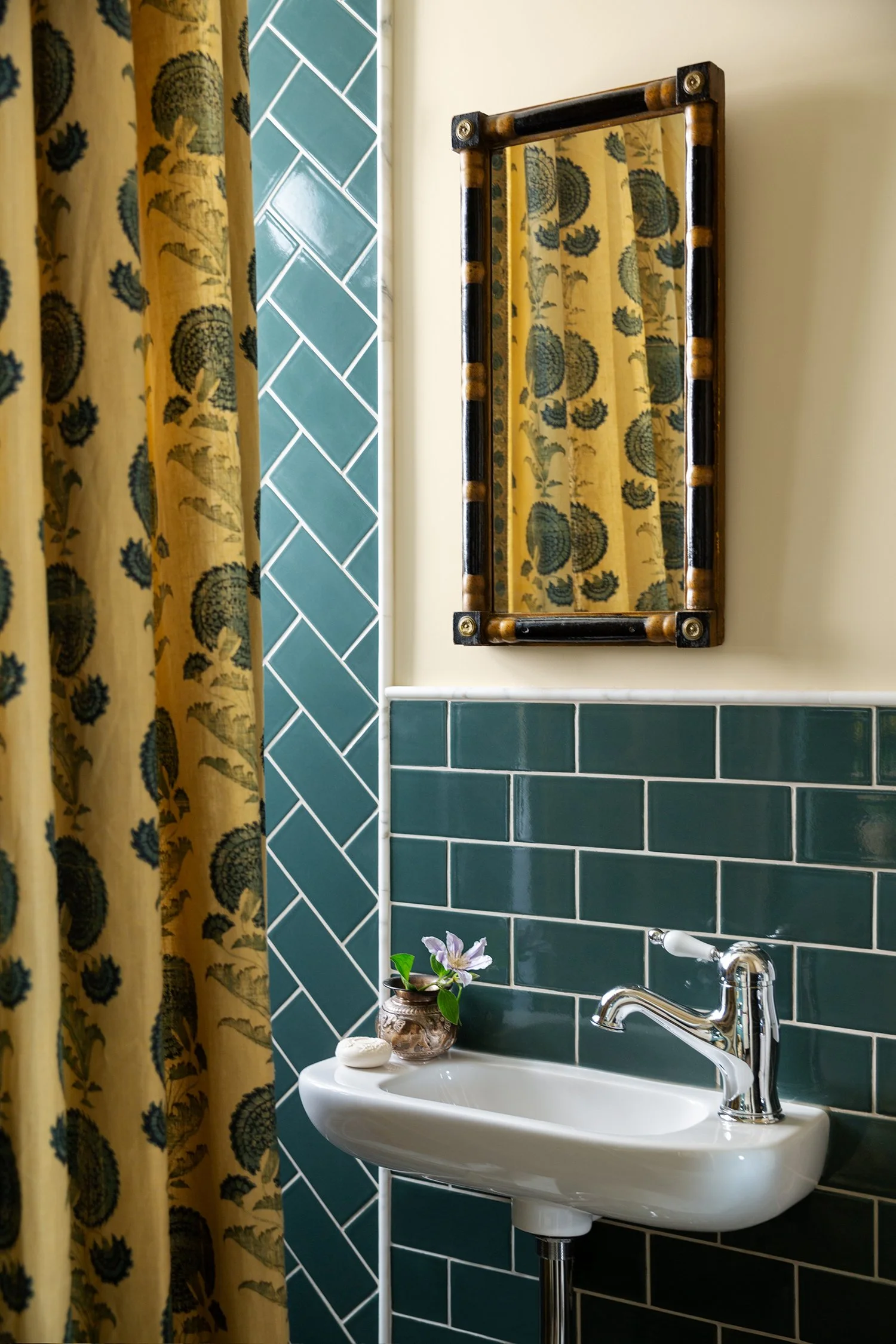 A small bathroom sink with a chrome faucet, a small vase with a flower, and soap on it. Hanging above is a wooden-framed mirror. The wall behind the sink features dark green tiles, and to the left, yellow curtains with a black floral pattern.