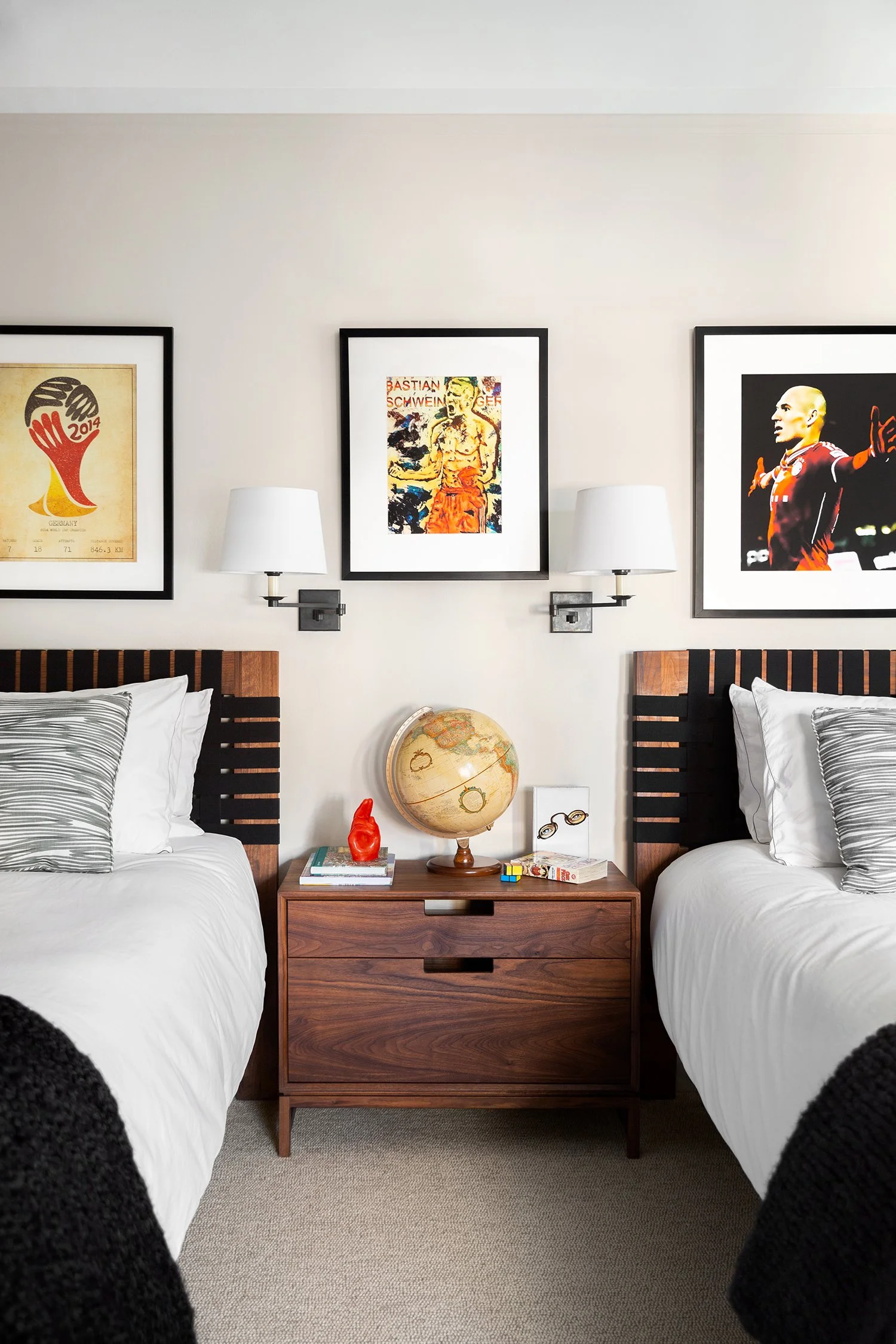 Twin beds with black headboards and white bedding, a wooden nightstand with a globe, glasses, a red sculpture, books, and a Rubik's cube, and wall-mounted lamps between framed artwork above.