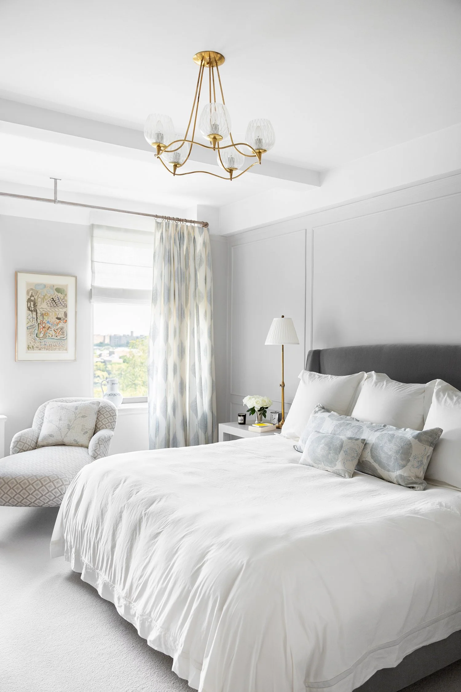 A bright, white bedroom with a chandelier hanging from the ceiling, a large bed with white bedding and pillows, a nightstand with a lamp and flowers, a window with patterned curtains, and an armchair near the window.