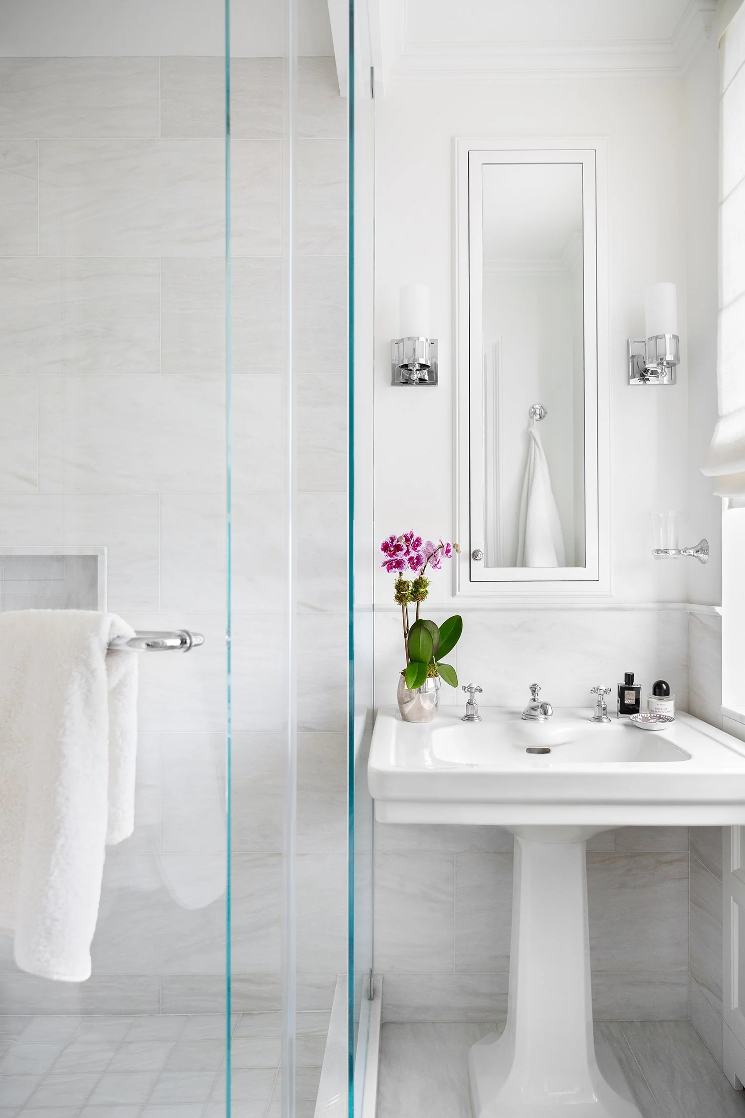 A minimalist, white bathroom featuring a porcelain pedestal sink with silver fixtures, a small potted orchid on the sink, a mirror with white framing, wall-mounted light fixtures, a towel hanging on a chrome bar, and a partially visible glass shower 