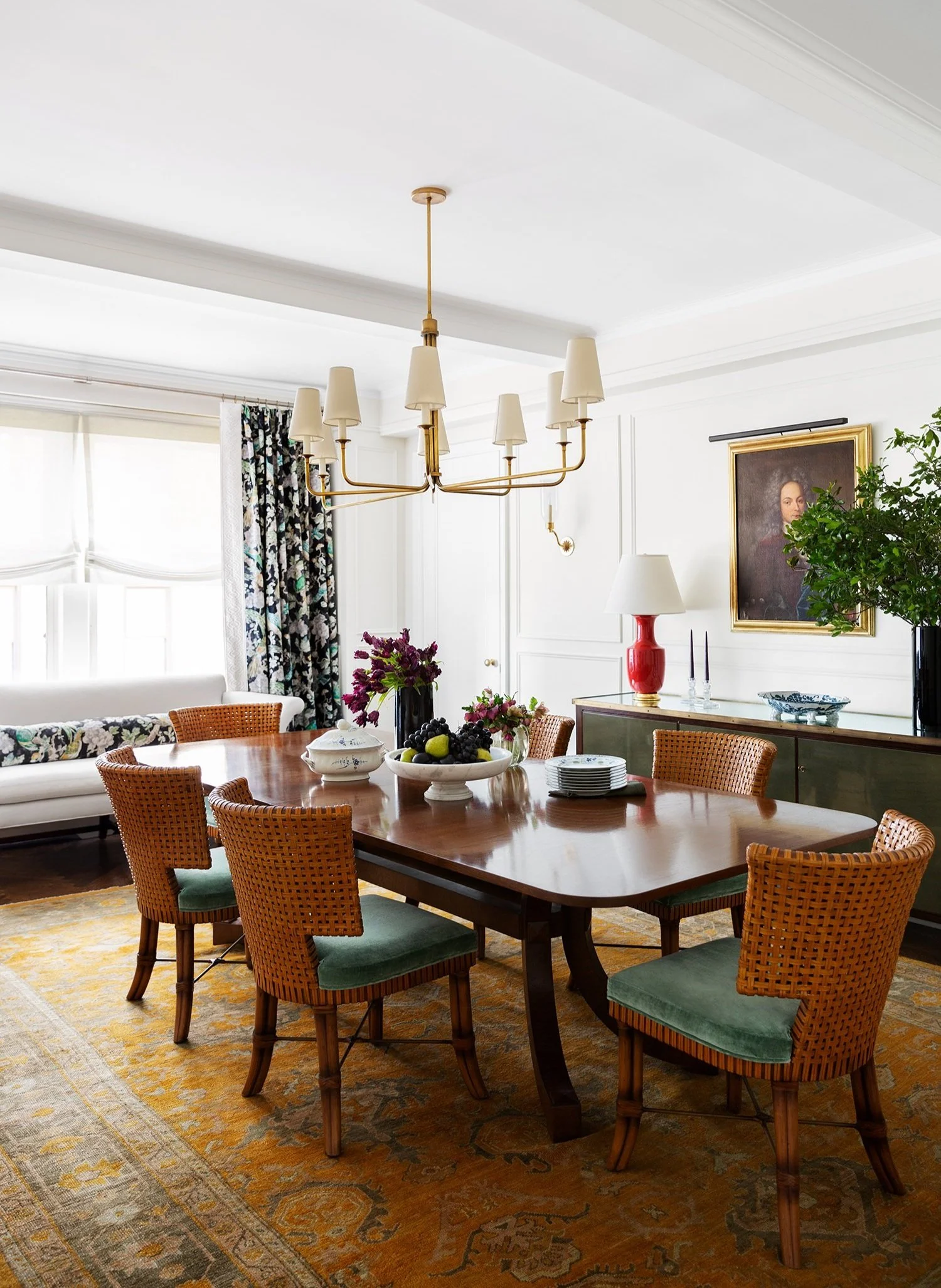 A dining room with a wooden table, surrounded by green cushioned wicker chairs. There is a chandelier hanging from the ceiling, and a sideboard with a red lamp, a large vase with greenery, picture frames, and a portrait hanging above it. The room has