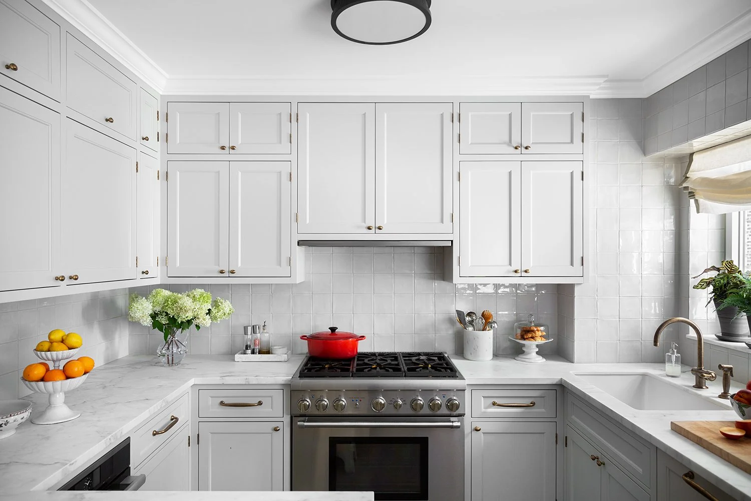 A clean, modern kitchen with white cabinets, a white marble countertop, a red pot on a stainless steel stove, and a window with a beige curtain, decorated with a small potted plant and fruit.