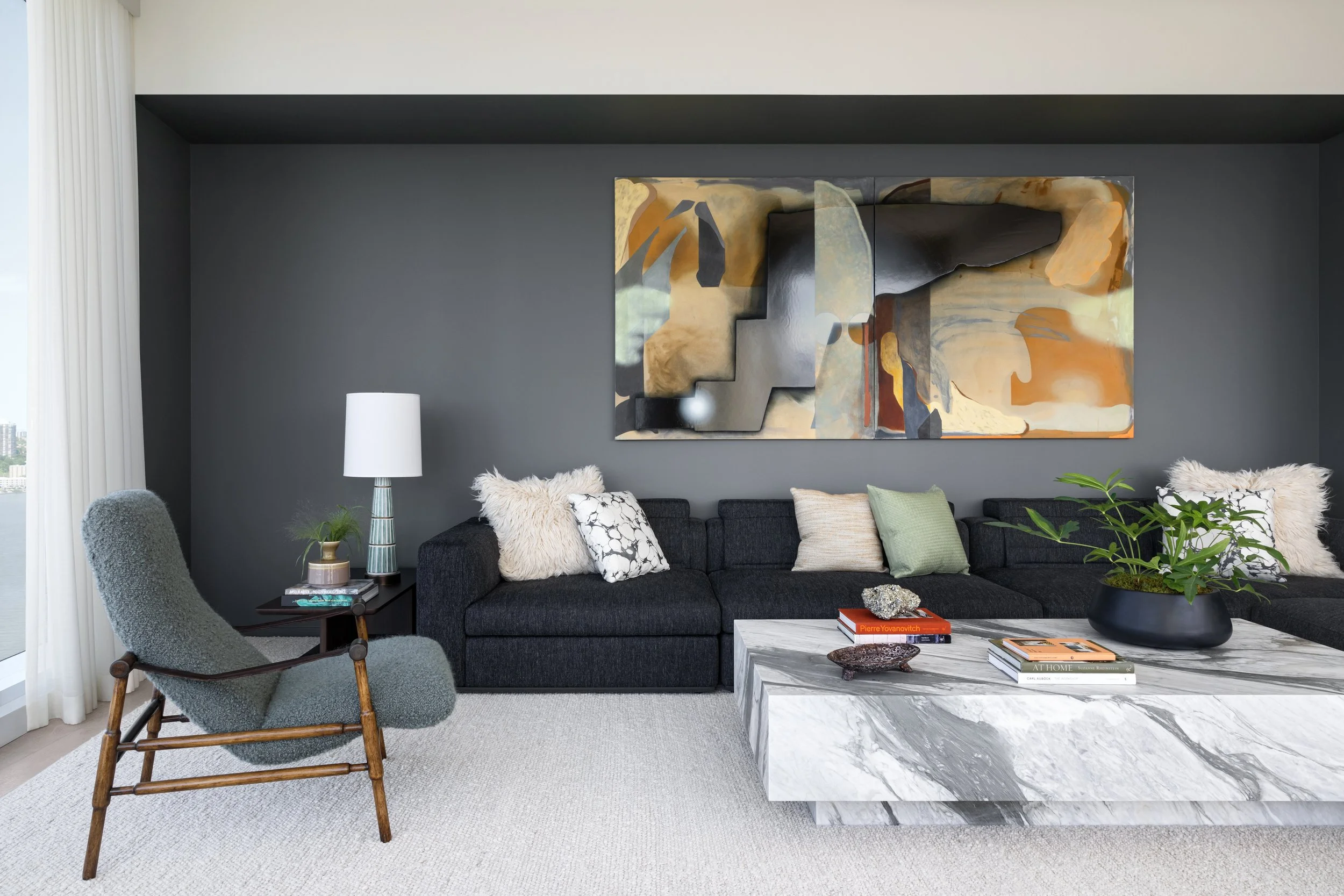 Modern living room with dark gray accent wall, abstract artwork above a black sectional sofa with various textured cushions, a marble coffee table with books and decorative objects, a gray armchair, a side table with a lamp and plants, and large wind