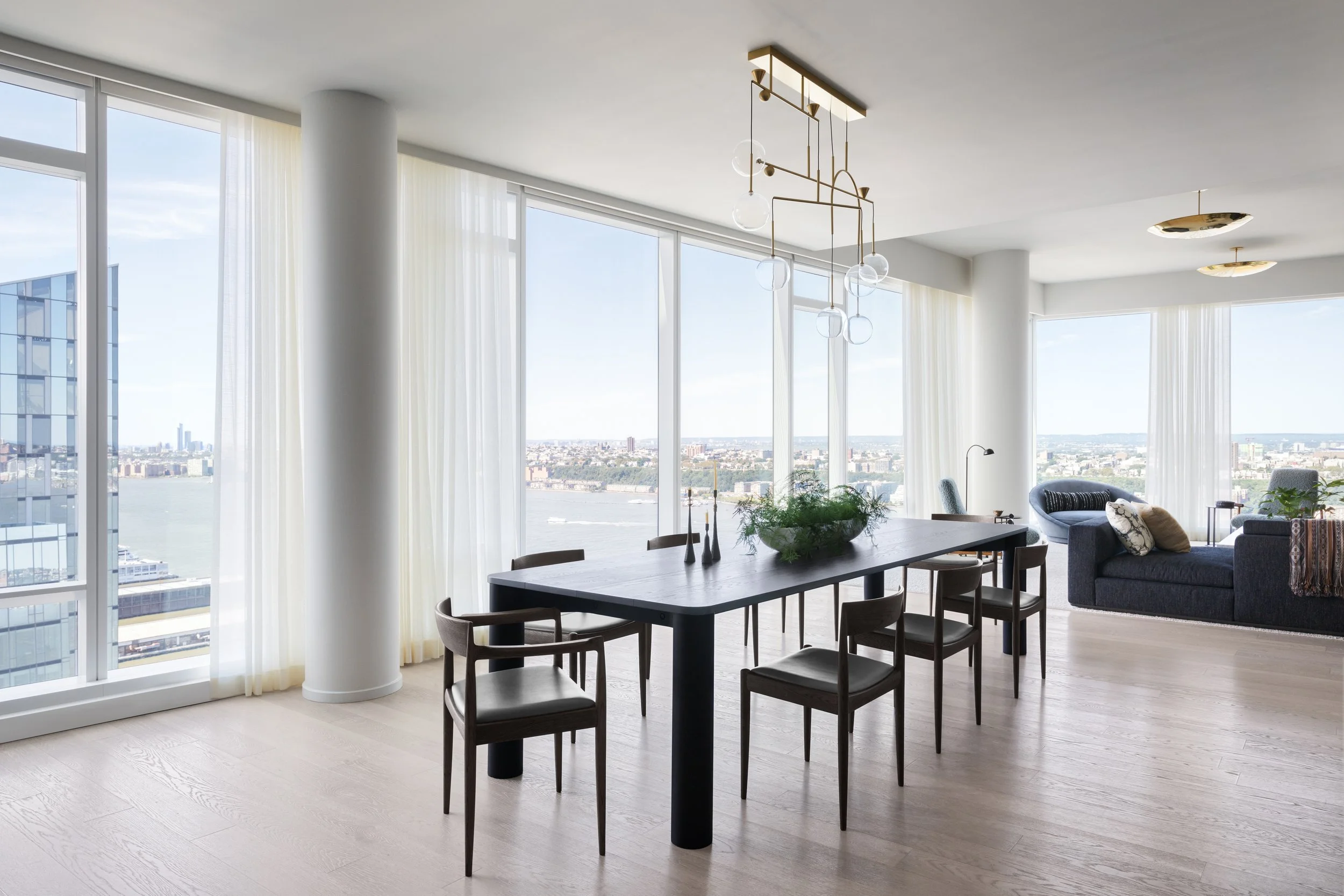 Modern high-rise apartment living room with floor-to-ceiling windows, a black dining table with chairs, a blue sofa with pillows, and minimalistic decor including a pendant light and plants, overlooking a cityscape and river.