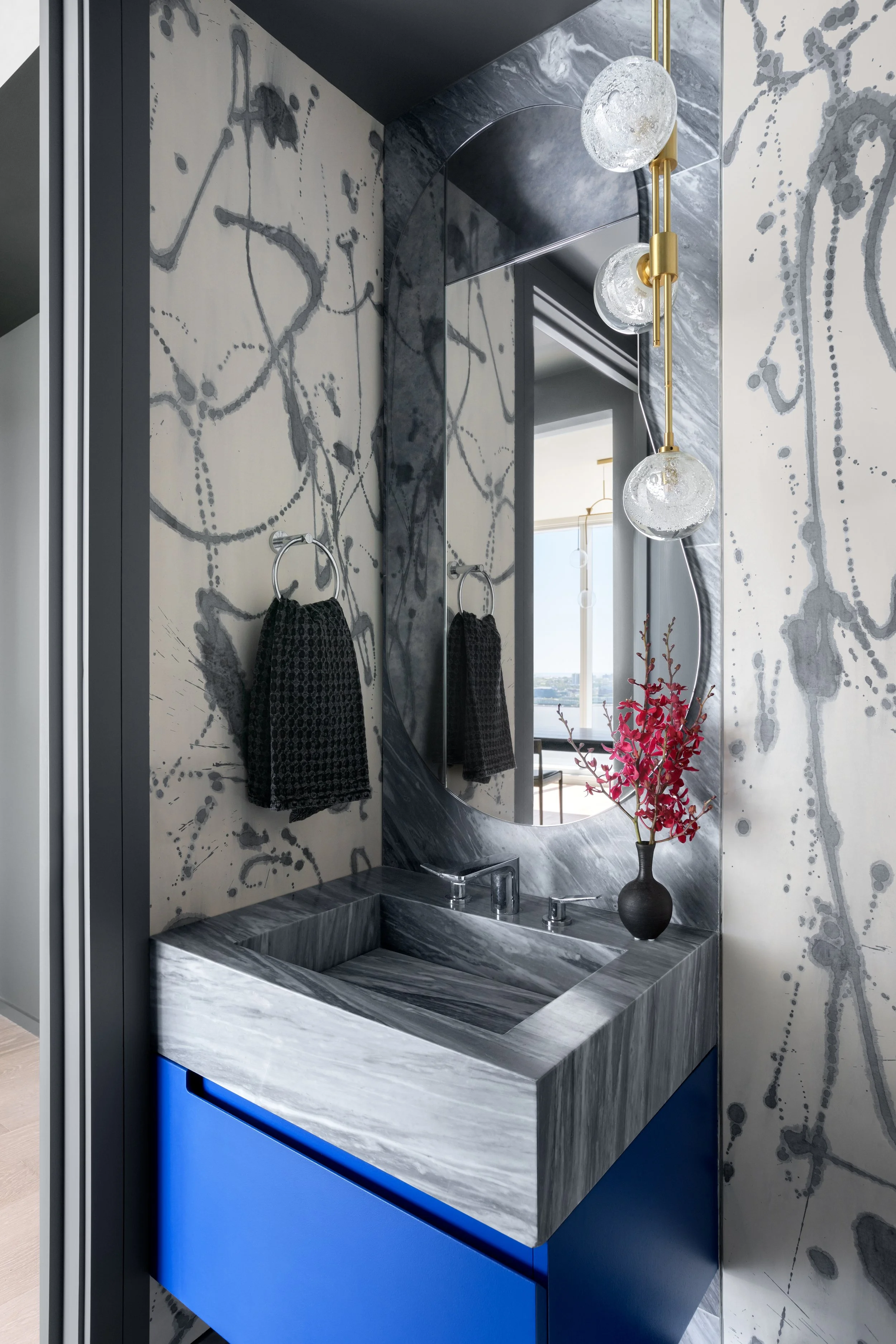 Modern bathroom vanity with a marble surface and blue cabinet, black vase with pink flowers, large round mirror, black towels, and a contemporary hanging light fixture with glass globes.