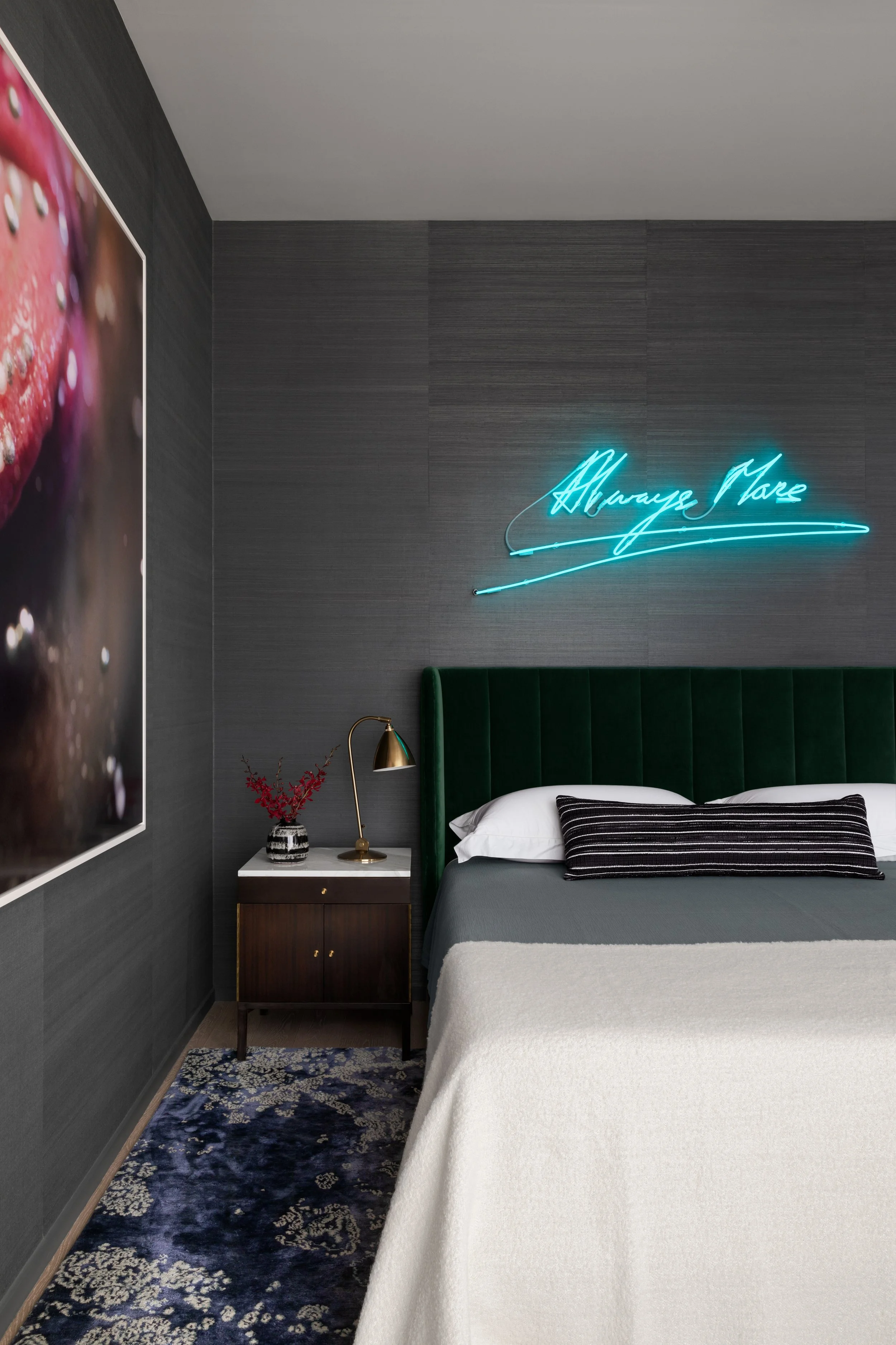 A bedroom with dark gray walls, a neon blue sign reading 'Always Be', a green headboard, a white bedside table with a gold lamp and a black and white decorative object, a dark patterned carpet, and a bed with white and dark striped pillows.