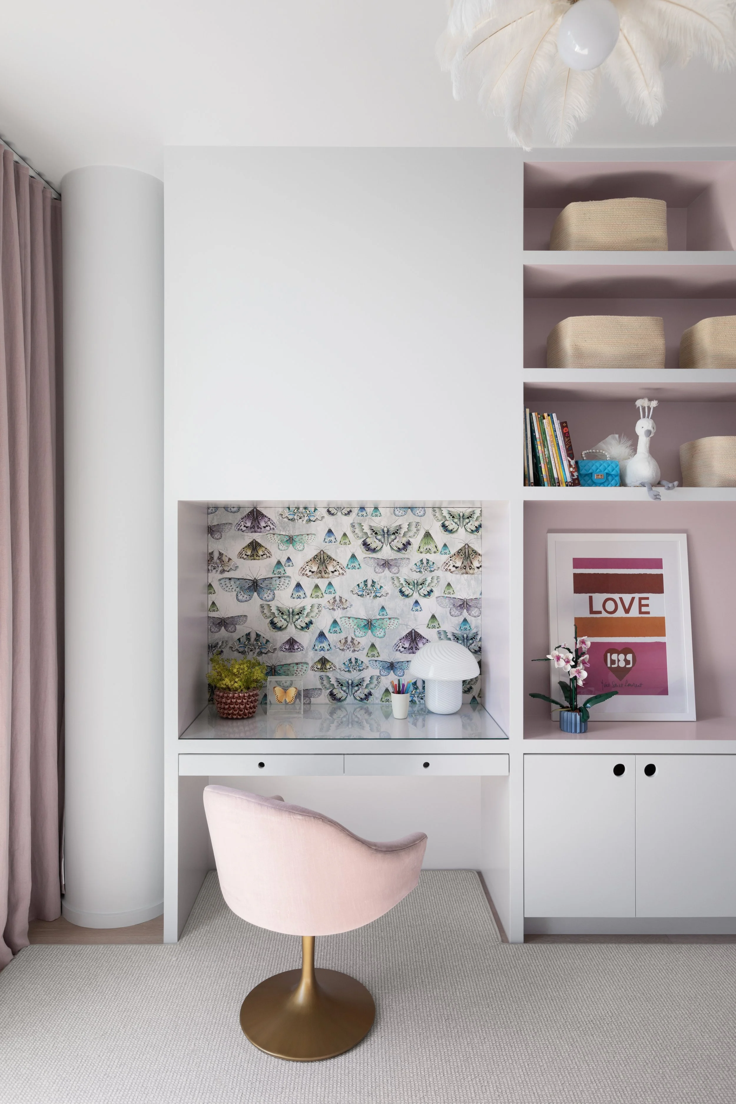 A cozy workspace with a white desk and pink chair in front, decorated with butterfly-patterned wallpaper or art background, a pink and white color scheme, books and art on the shelves, a framed quote that says "LOVE," and a decorative plant.