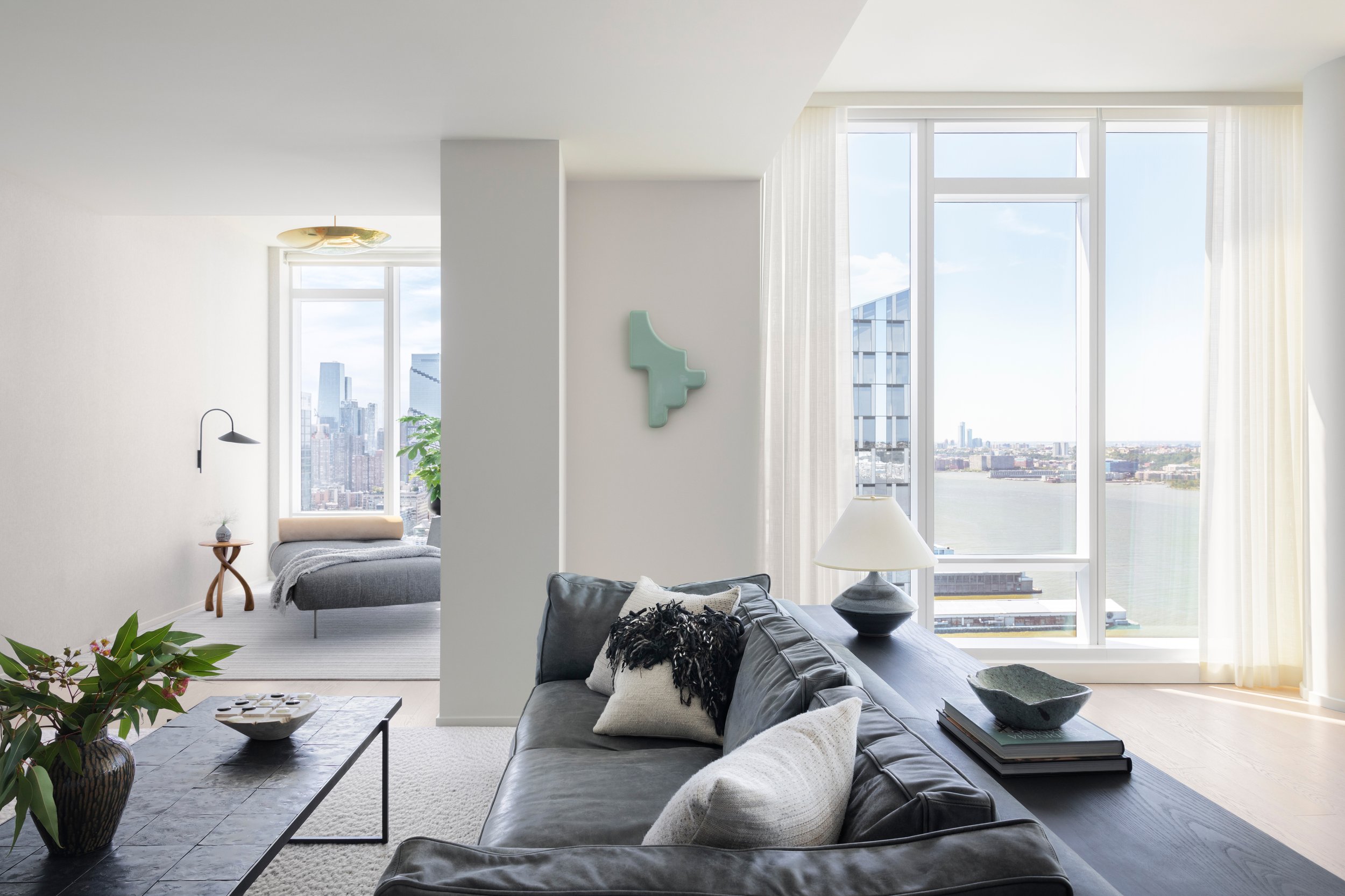 Modern living room with large windows overlooking a city skyline and water view, decorated with a black leather sofa, white pillows, a glass lamp, and a black coffee table.