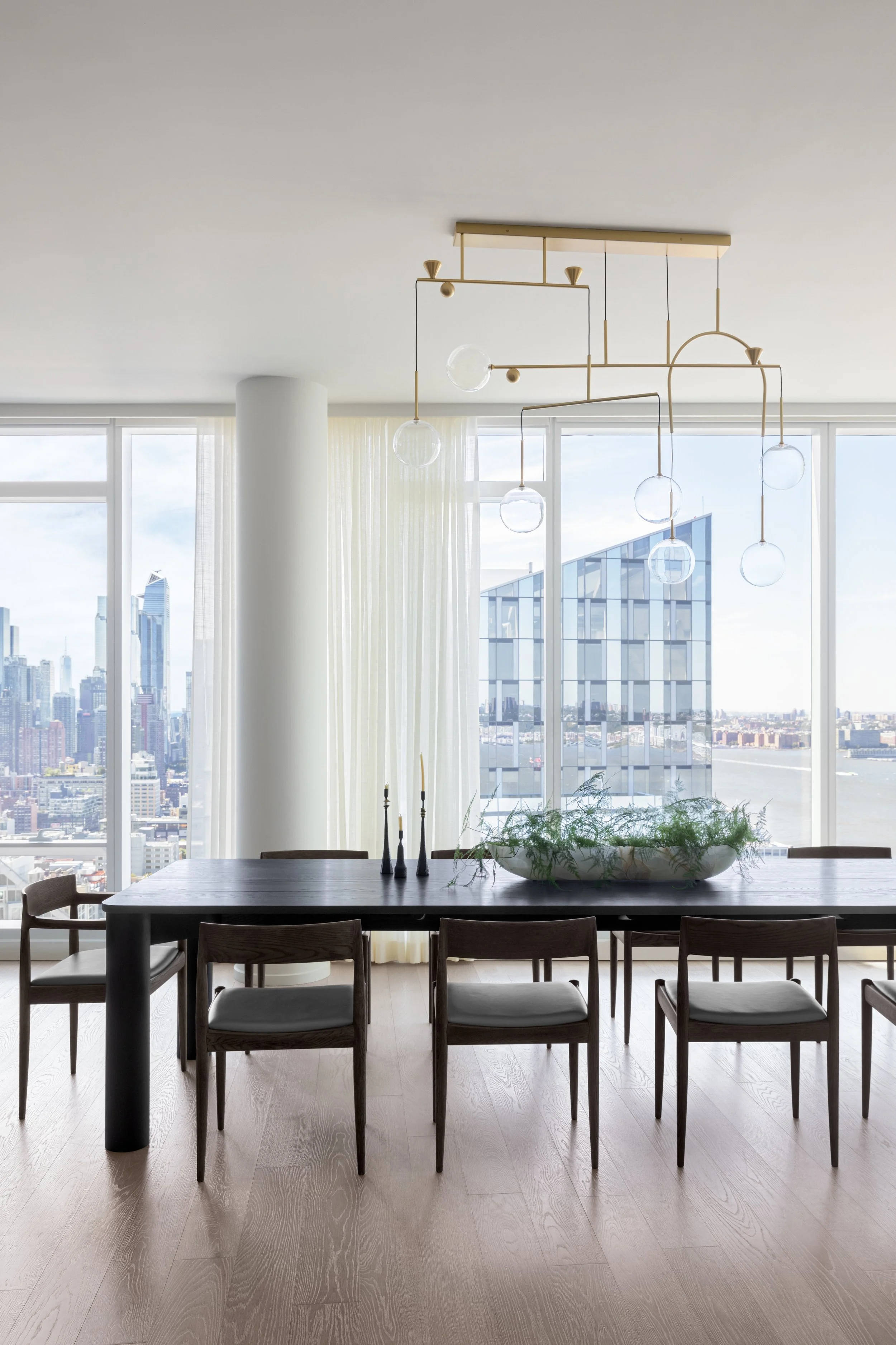 Modern dining room with a black table, six chairs, a large glass bowl with green plants, three black candles, a unique chandelier with globe lights, and large windows showing city skyscrapers and water outside.