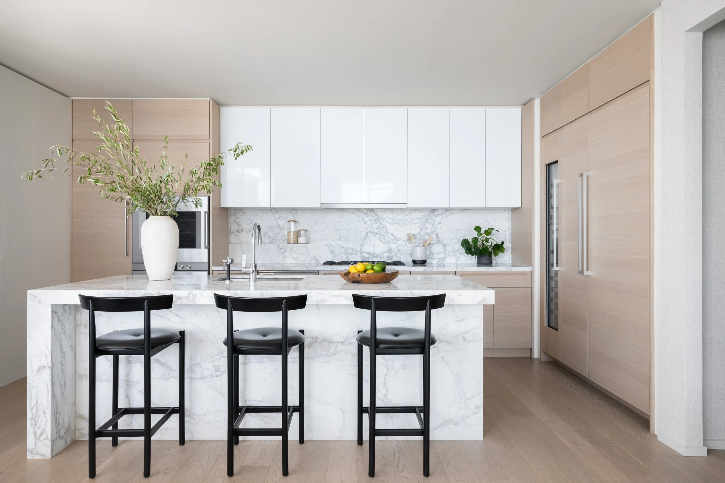 Modern kitchen with white and light wood cabinets, marble island, black stools, plants, and minimalist decor.