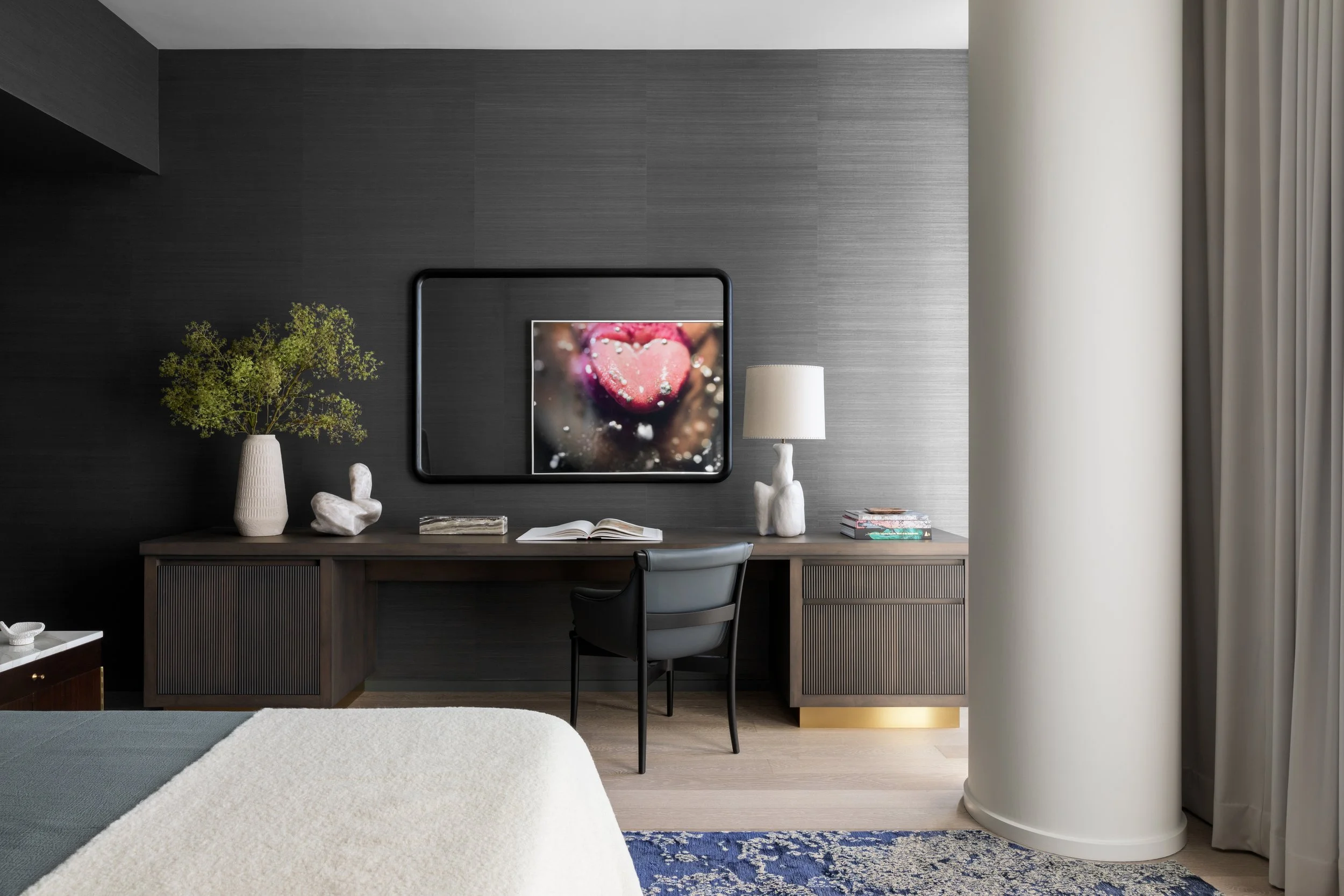 Modern hotel or office workspace with a dark wood desk, decorative vases, a white table lamp, open books, and a black chair against a dark textured wall, near large curtains.