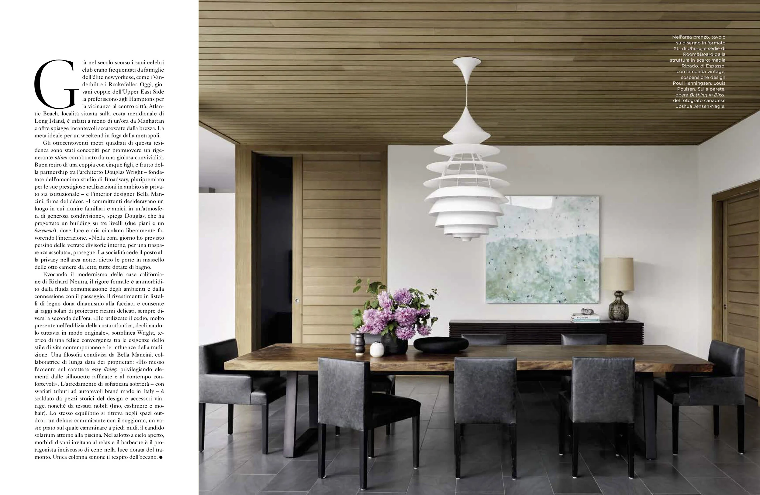 Modern dining room with wooden ceiling and a large wooden table surrounded by six black and gray chairs. A white, layered pendant light hangs above. A black sideboard with a stack of books, a table lamp, and decorative items, and a door with a wooden
