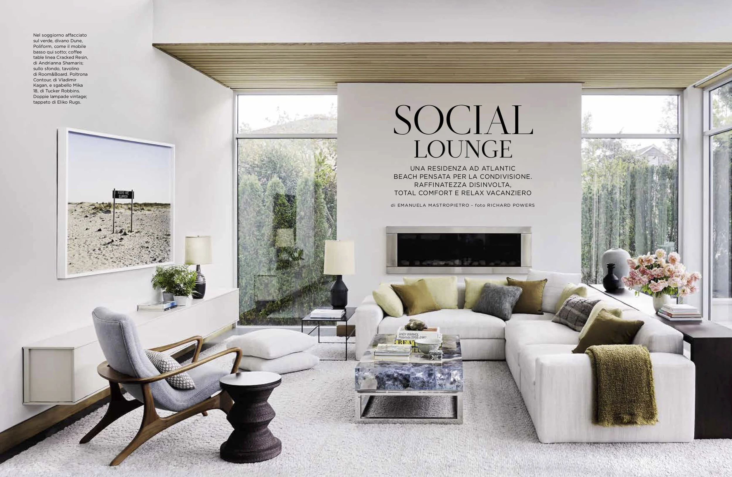 A modern living room with a white sectional sofa, throw pillows, a marble coffee table, an armchair, side tables with lamps, a fireplace, large windows, and wall art. The room is decorated with plants, books, and flowers.