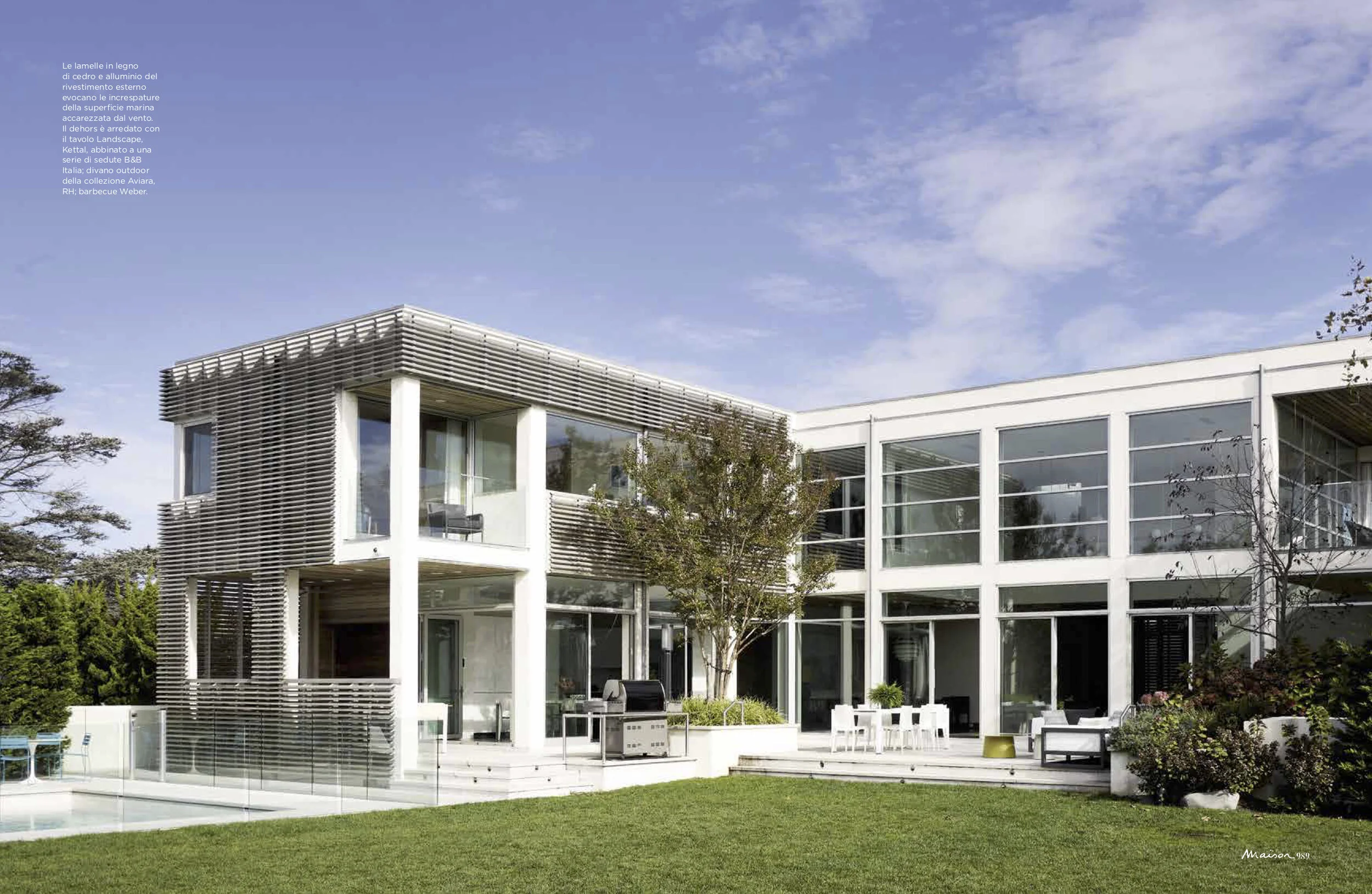 Modern two-story house with large glass windows, featuring wooden slats on part of the exterior, surrounded by a lush green lawn and outdoor furniture, with a partly cloudy sky overhead.