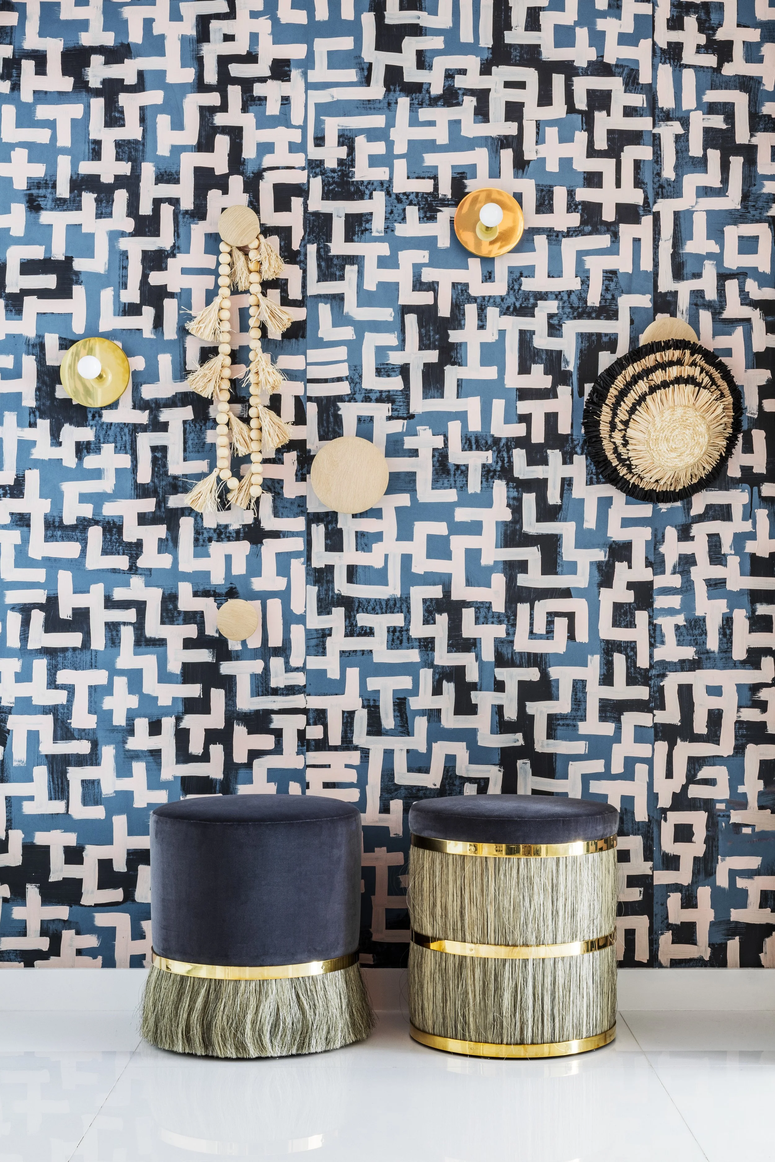 A decorative wall with a geometric patterned wallpaper in blue, black, and pink, adorned with various wall hangings, including a shell and tassel piece, two yellow circular wall decorations, and a woven basket. There are two upholstered stools with b