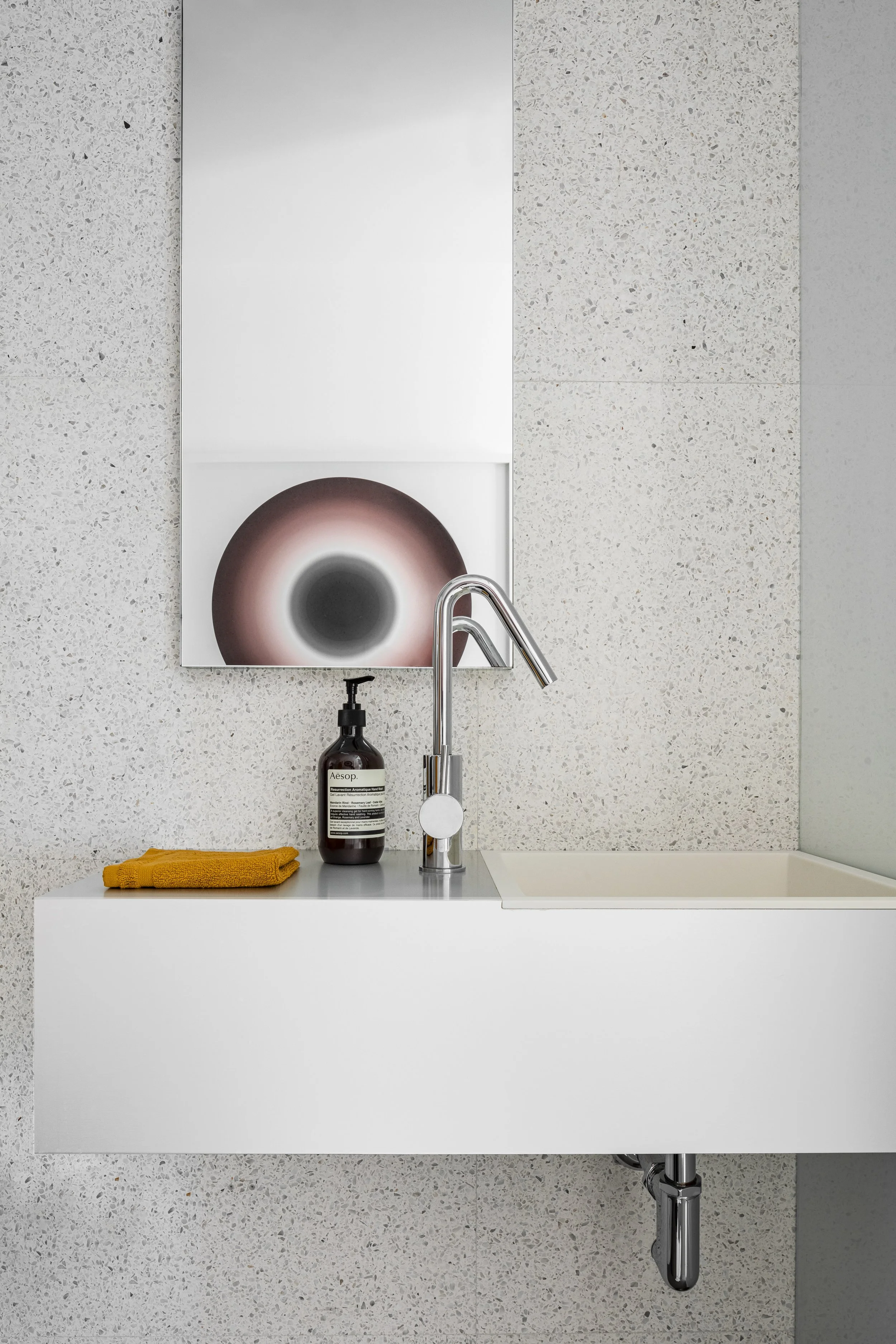 A modern bathroom sink with a white basin, chrome faucet, a black soap dispenser, and a yellow cloth on the countertop. A large abstract art piece featuring a circular design hangs above the sink, with textured light-colored walls surrounding it.