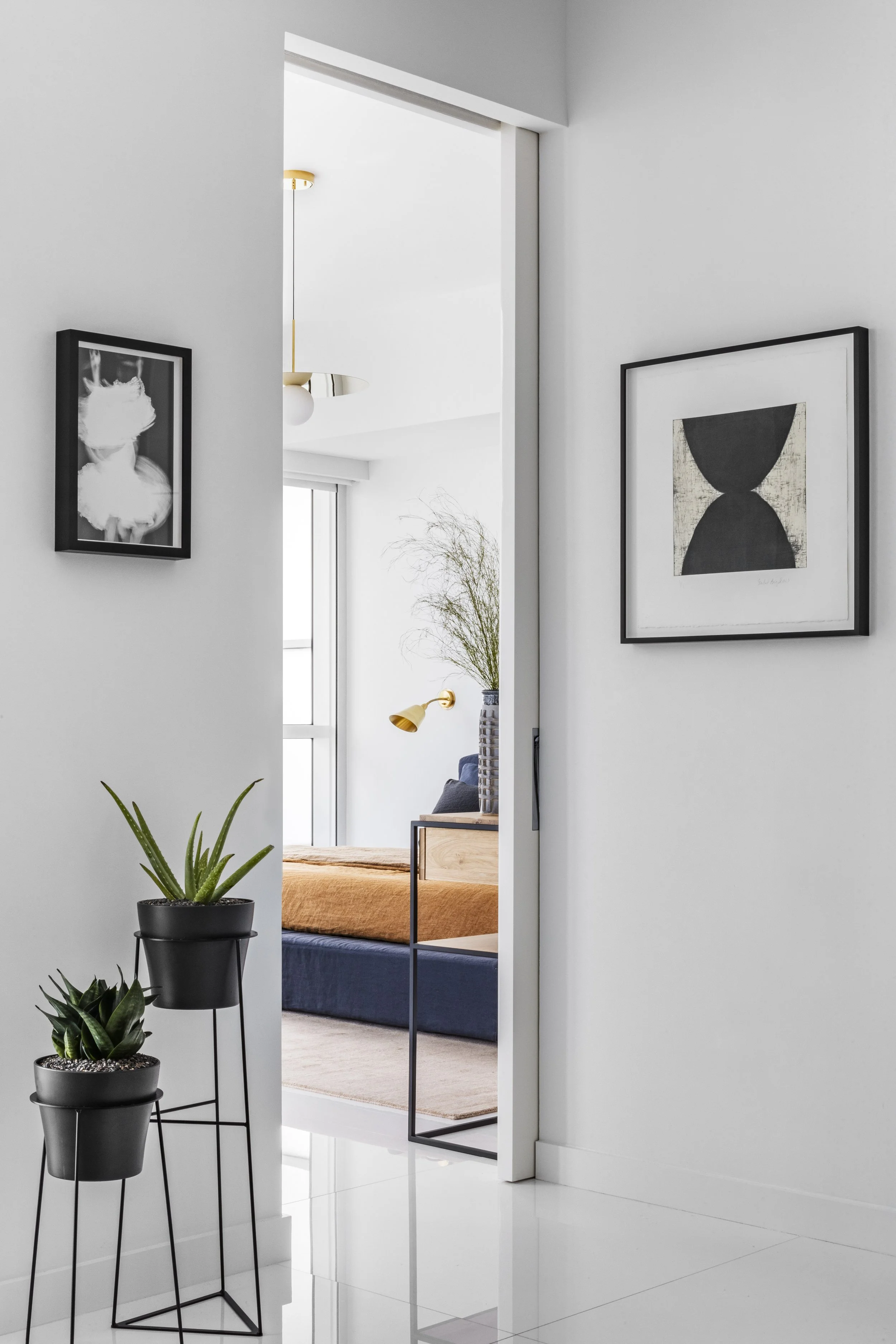 Interior view of a modern apartment hallway leading to a bedroom with a bed, a wooden nightstand, a large vase with tall plant, and a wall-mounted gold lamp. The hallway has two framed abstract art pieces and potted plants on two black metal stands.