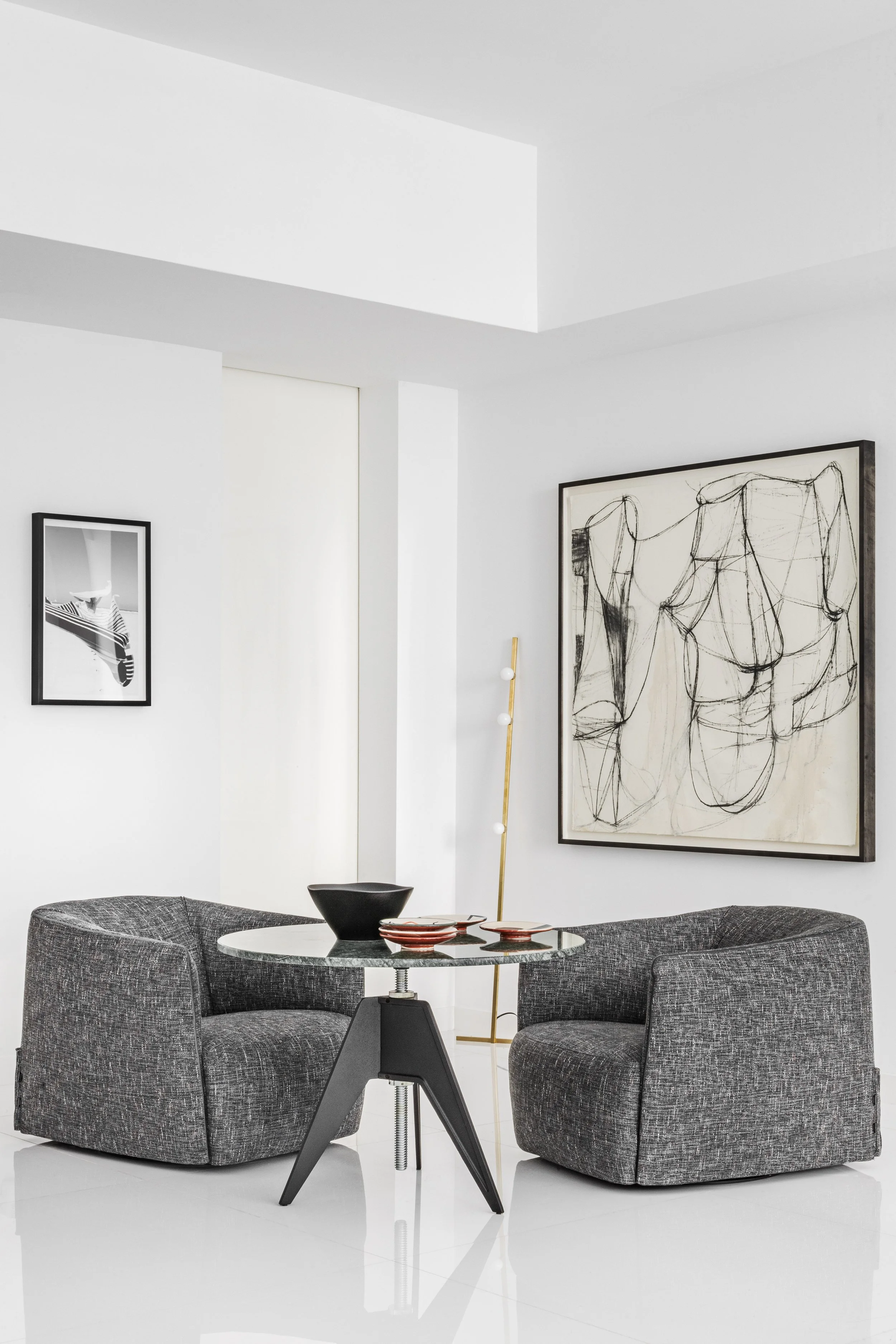 Modern living room with two gray upholstered armchairs, a round glass table with black bowl and red plates, large abstract black and white artwork, smaller black and white framed photo, and a tall gold floor lamp with white globes, all in a minimalis