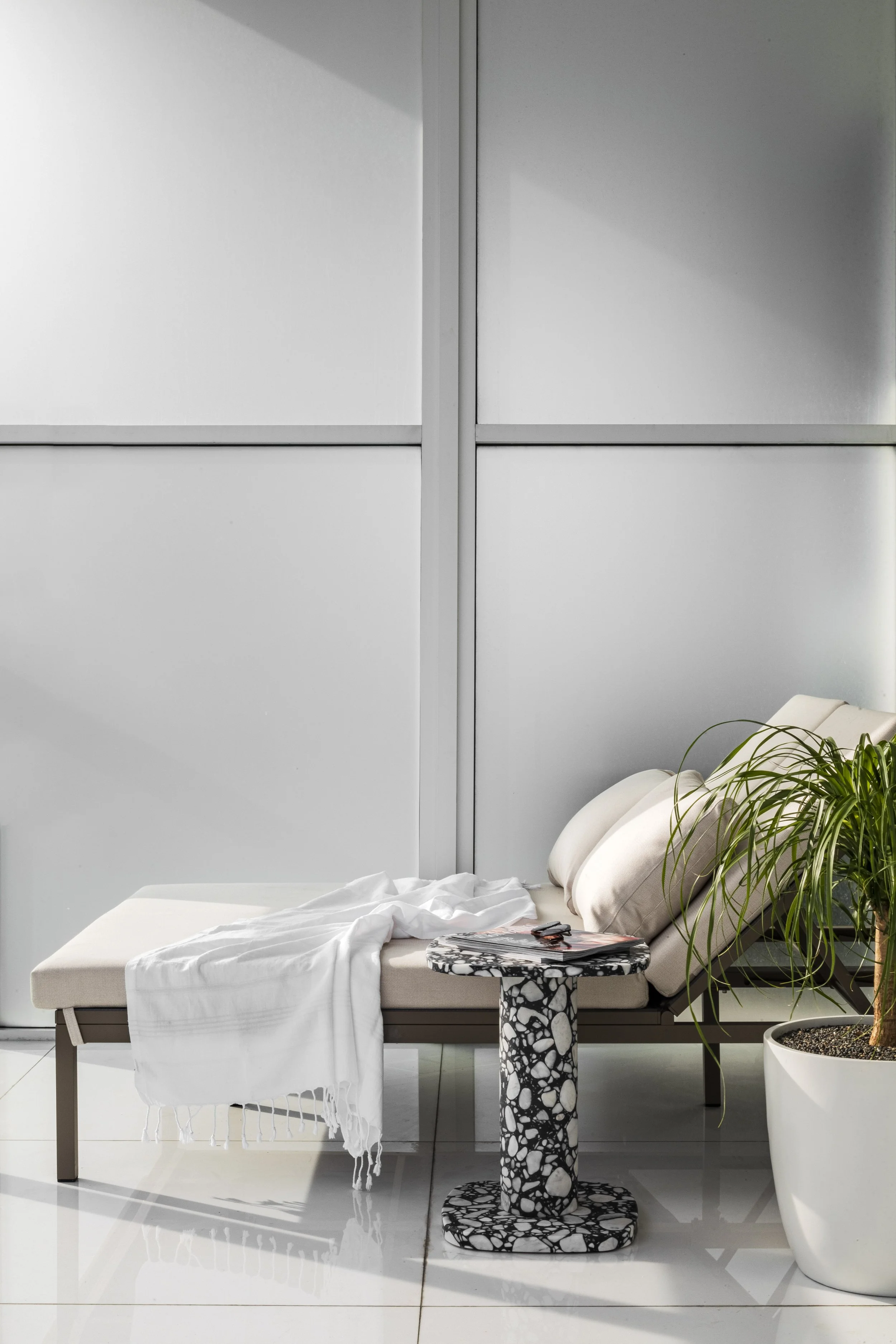 A modern medical or spa waiting area with a beige lounge chair, white blanket, a black and white terrazzo side table with magazines and sunglasses, green potted plant, white tiled floor, and frosted glass wall panels.