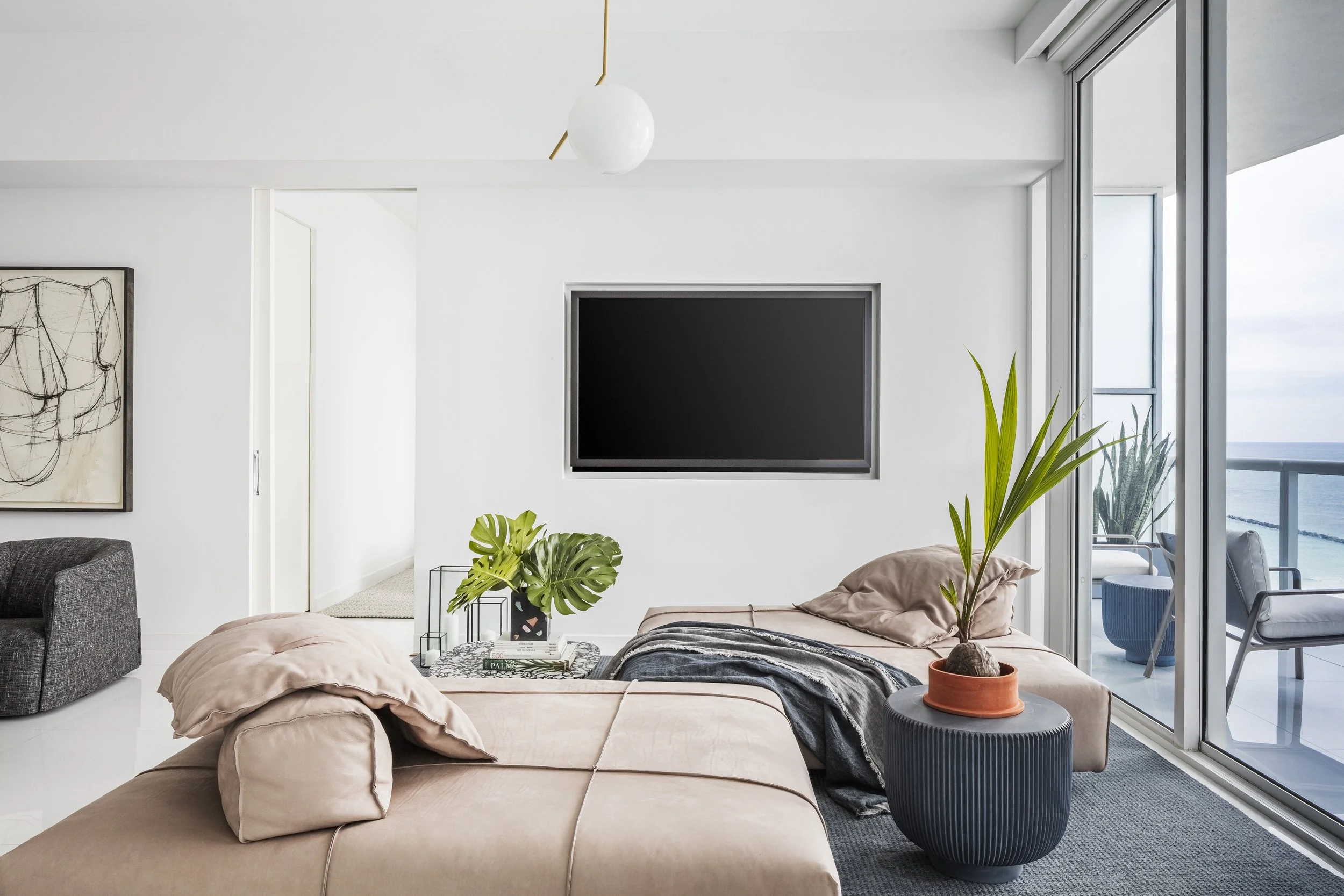 Modern living room with a beige sofa, potted plants, a wall-mounted TV, and a balcony with ocean view.