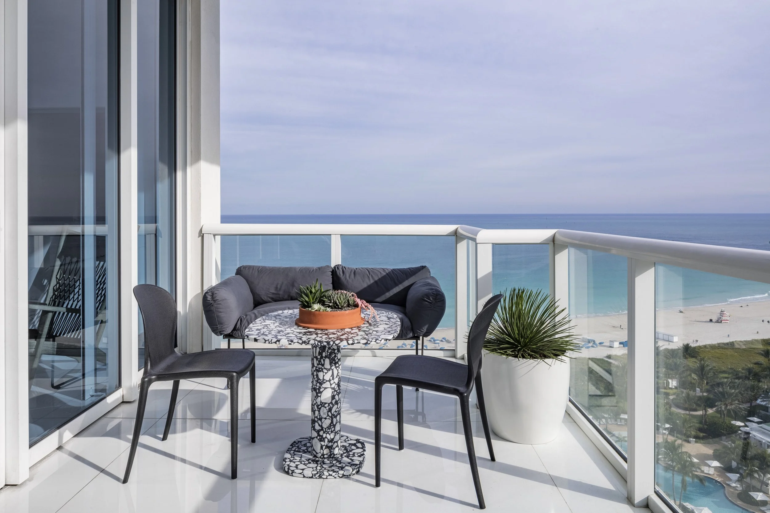 Balcony with black chairs, a round patterned table with potted plants, gray cushioned seating, large potted plant, overlooking beach and ocean under partly cloudy sky.